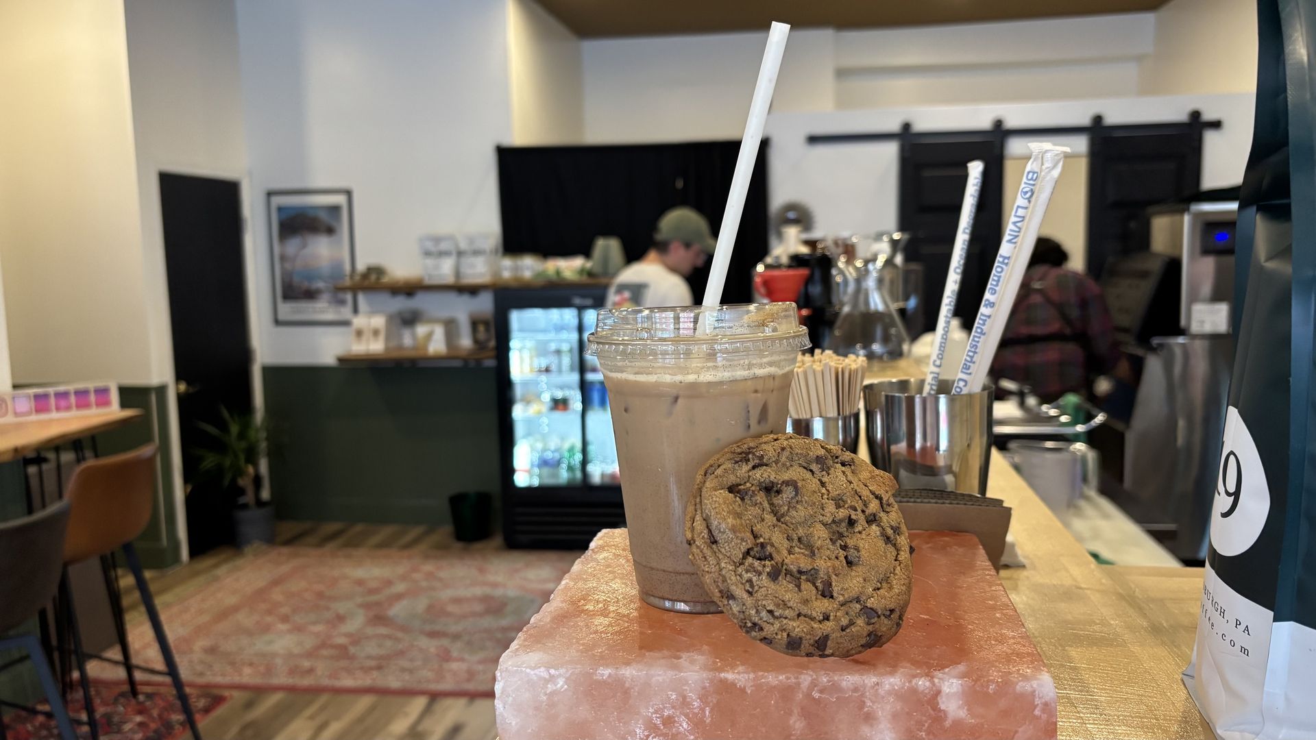 Iced coffee in a clear plastic cup with a white straw and a chocolate chip cookie resting on a pink salt block on a wooden counter in a cozy café interior.