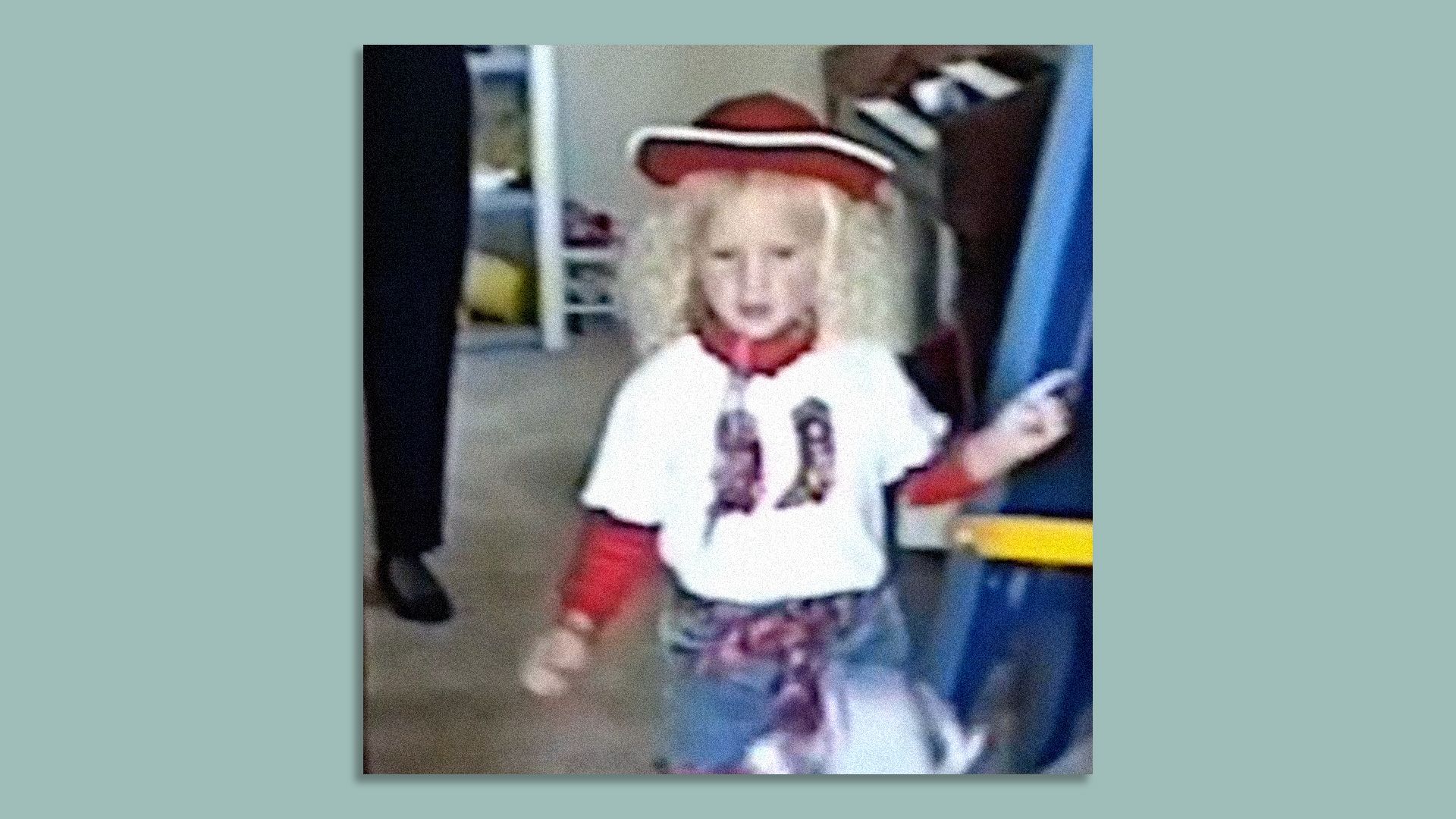 Blurry photo of a young child in a costume: white shirt, red scarf and sleeves, curly blond hair, wearing a red hat with white trim, standing indoors near a blue and yellow play structure.