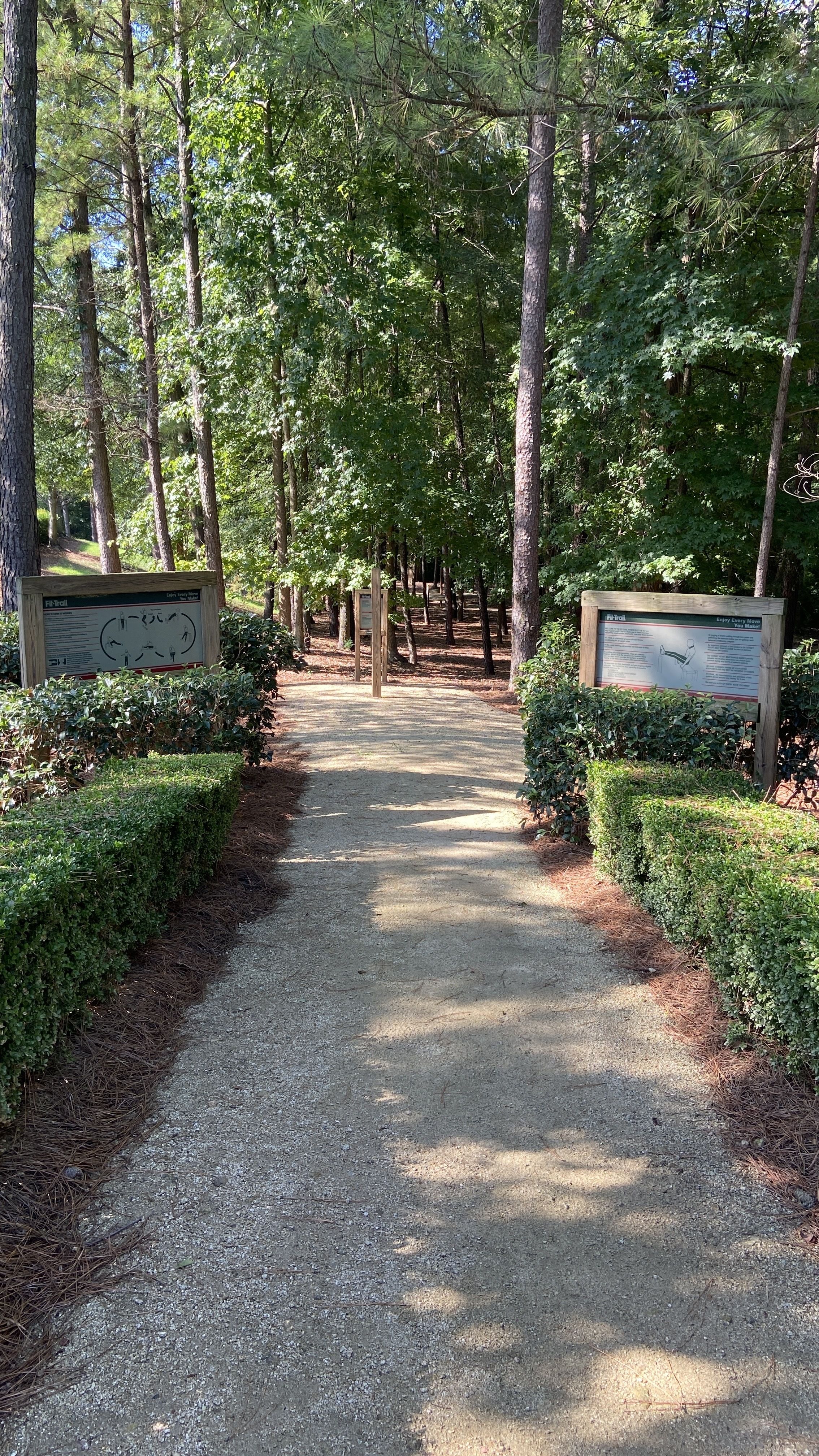 Gravel path through a green forested area with trimmed hedges and two wooden signboards on either side providing information about the FiTrail fitness trail.