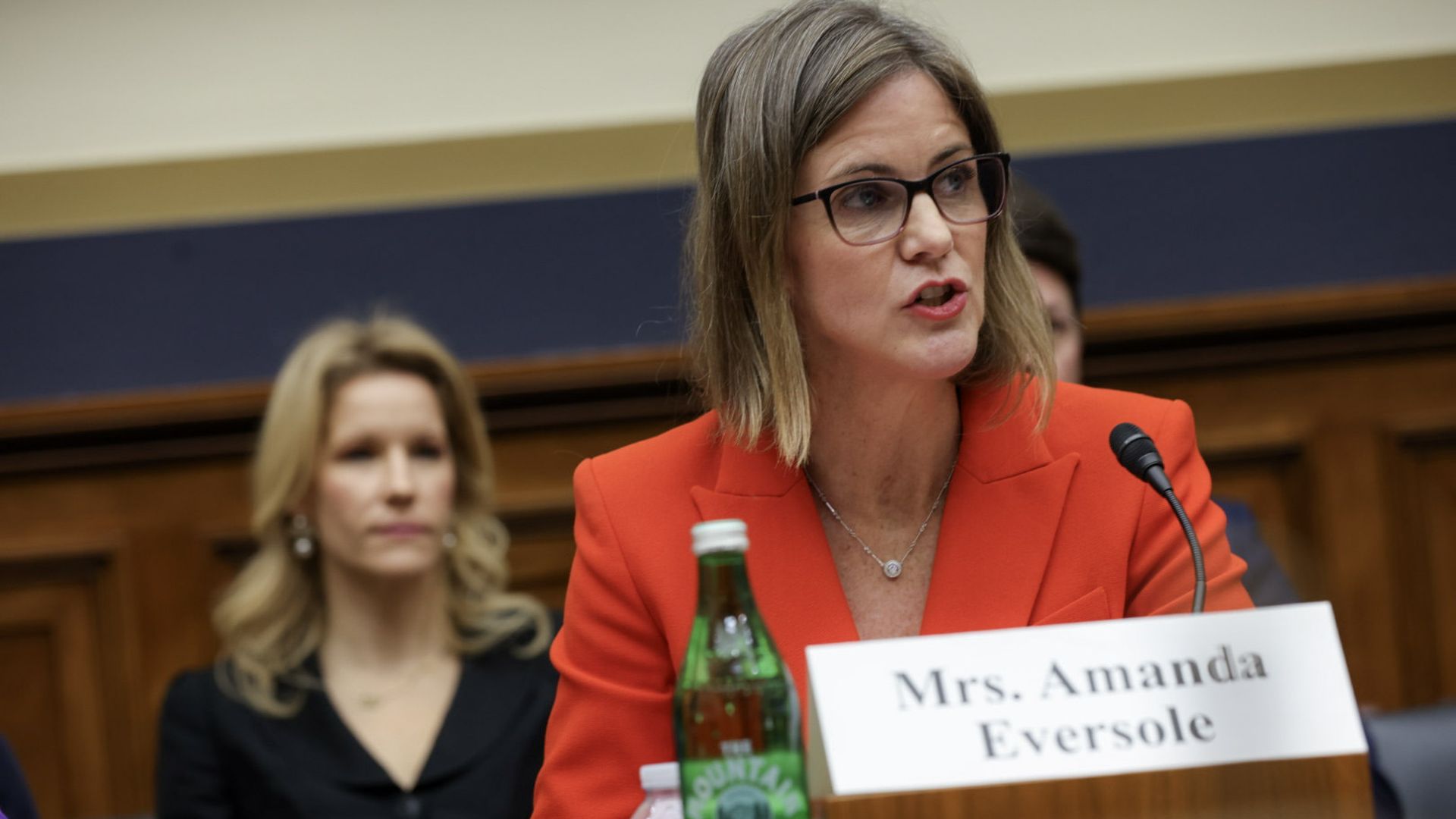 Woman in red blazer speaking at a microphone during a formal meeting, with a nameplate reading "Mrs. Amanda Eversole" and a green bottle of Mountain Valley water in front.