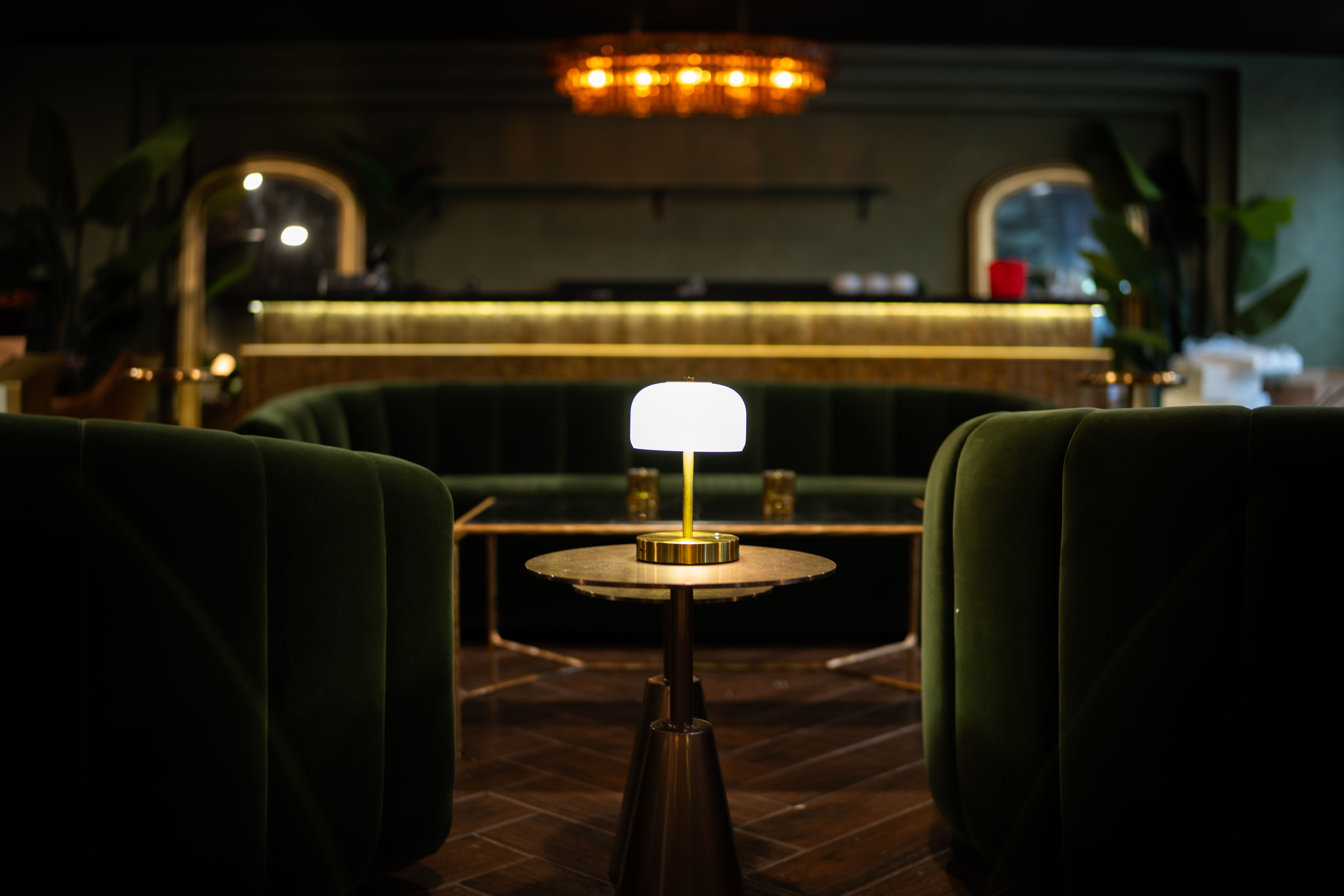 Dimly lit lounge with deep green velvet chairs, a small round table with a glowing white lamp, and a gold-accented bar with warm lighting in the background.