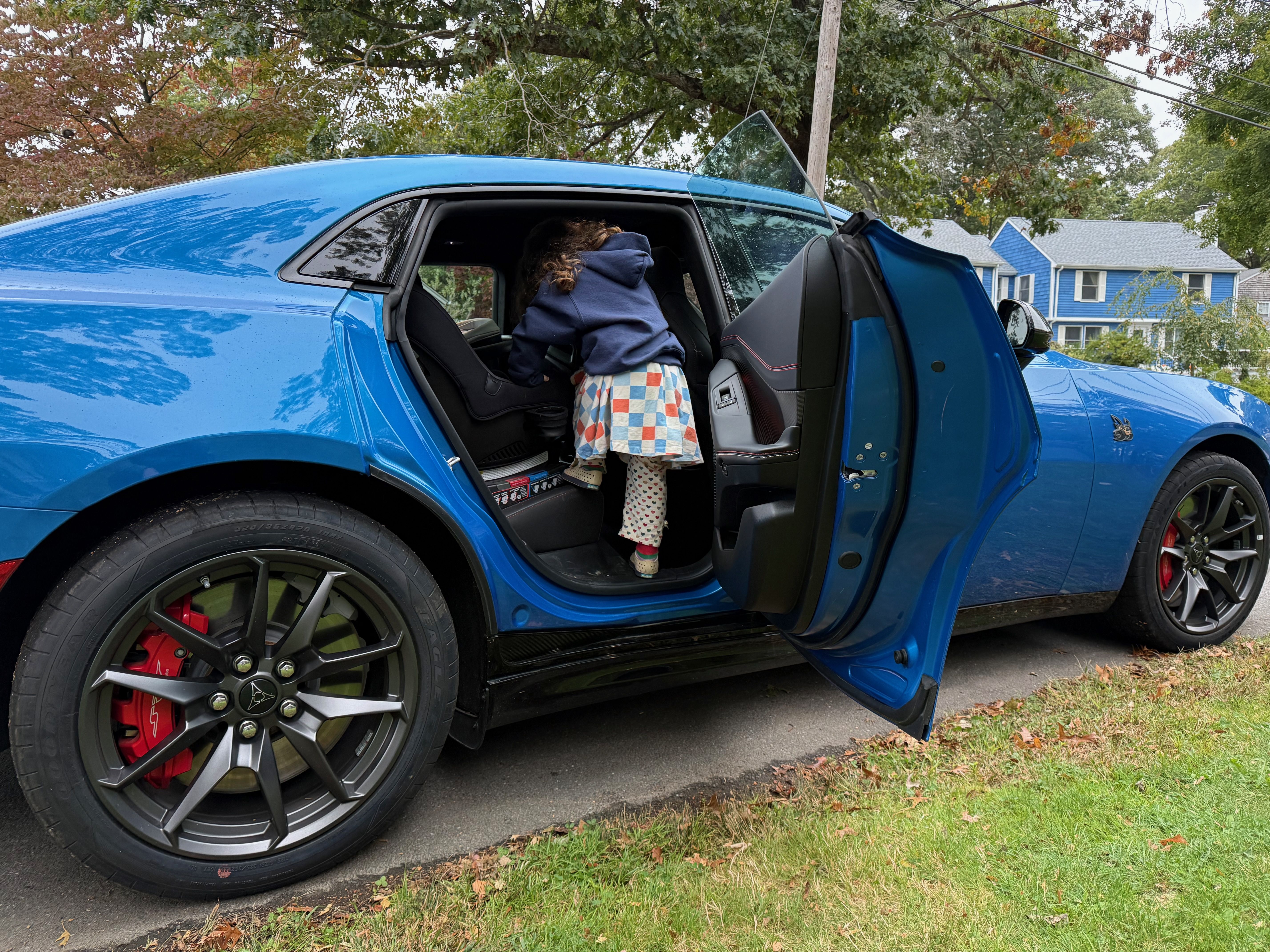 A 4-year-old climbs into a car seat in the back of a blue Dodge Charger EV, parked in a residential driveway.