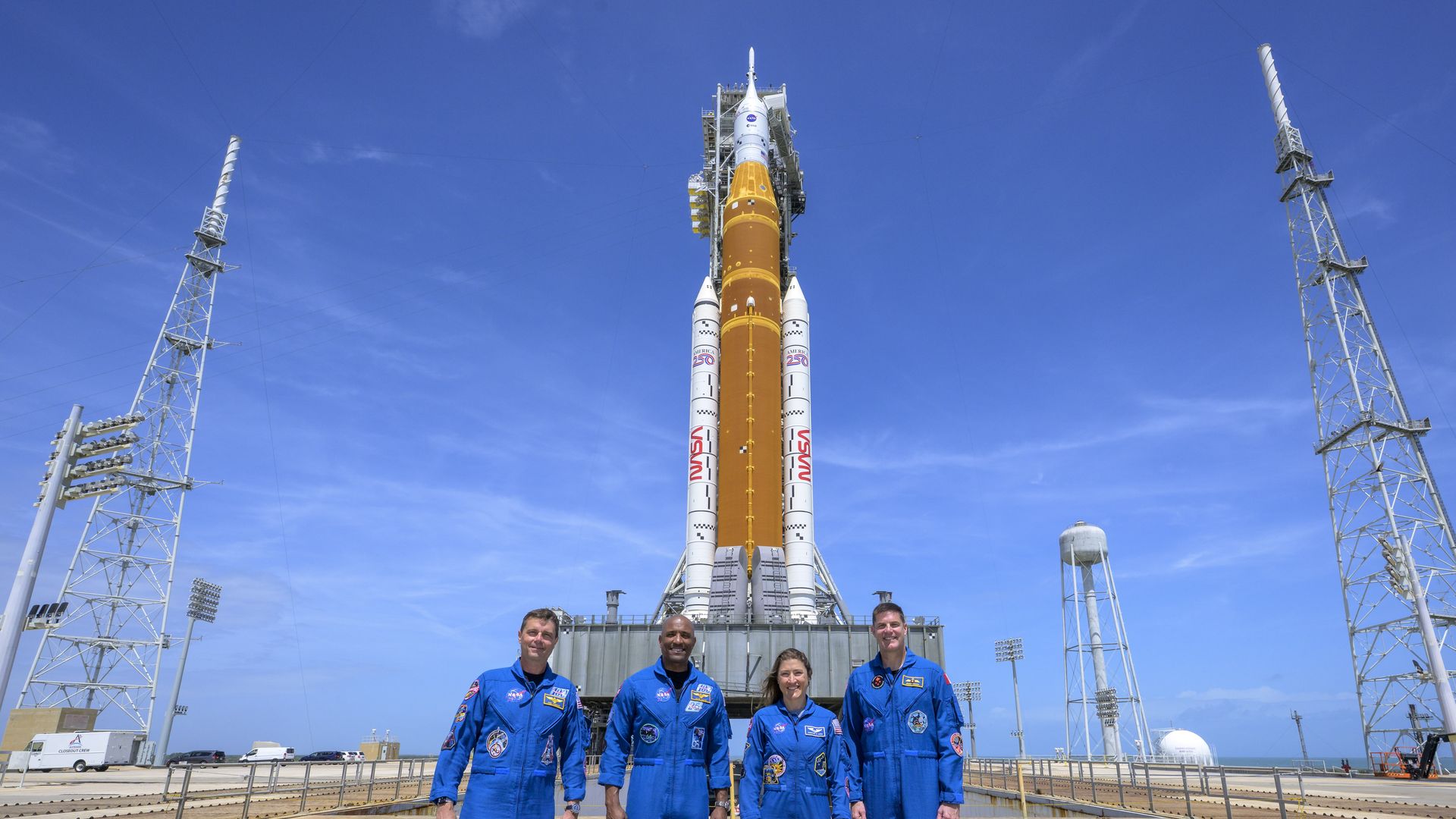 Image show four astronauts in blue flight suits in front of the large Artemis rocket.