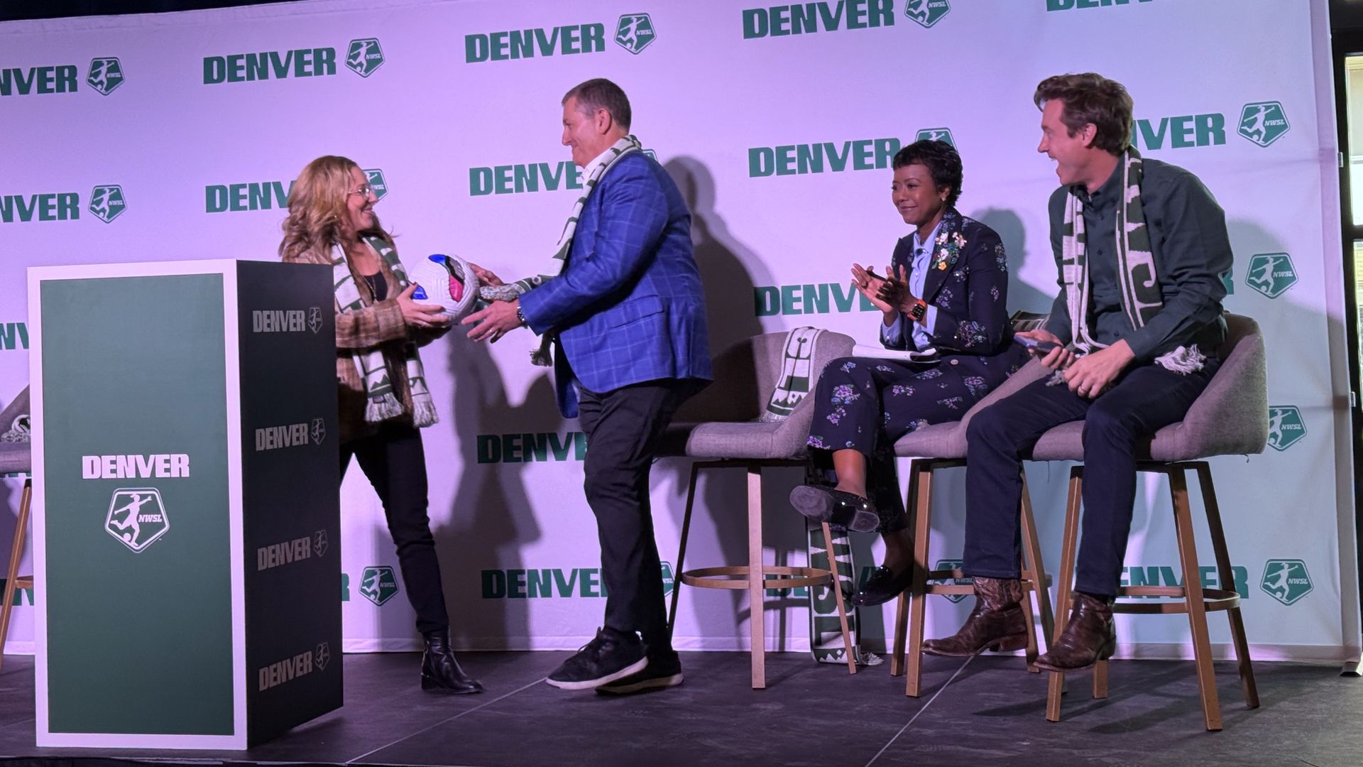 A woman hands over a white soccer ball to a man in a blue jacket while two other people look on. 