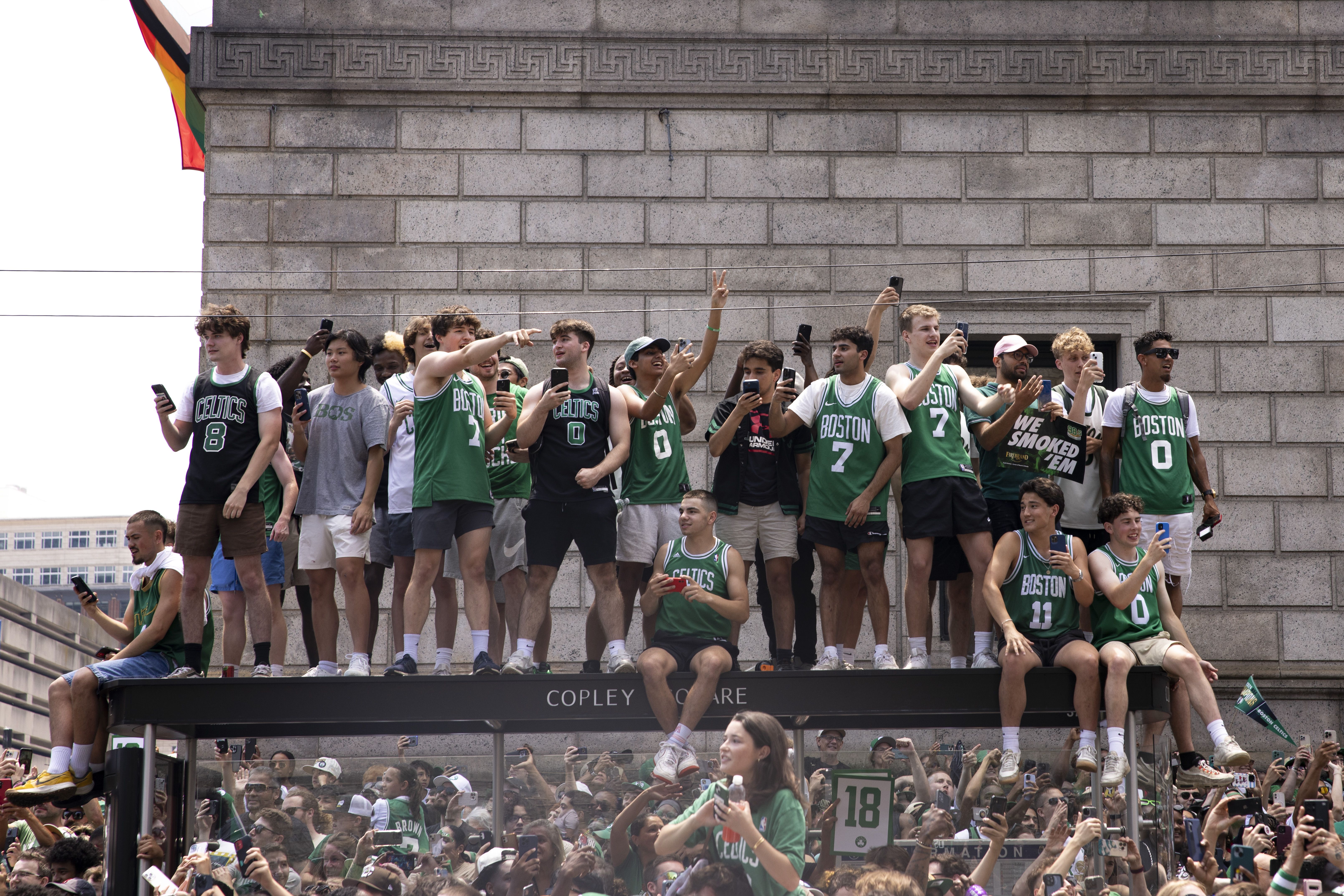 A group of young men with Celtics jerseys on stand on top of the Copley Square bus stop during the Celtics parade.