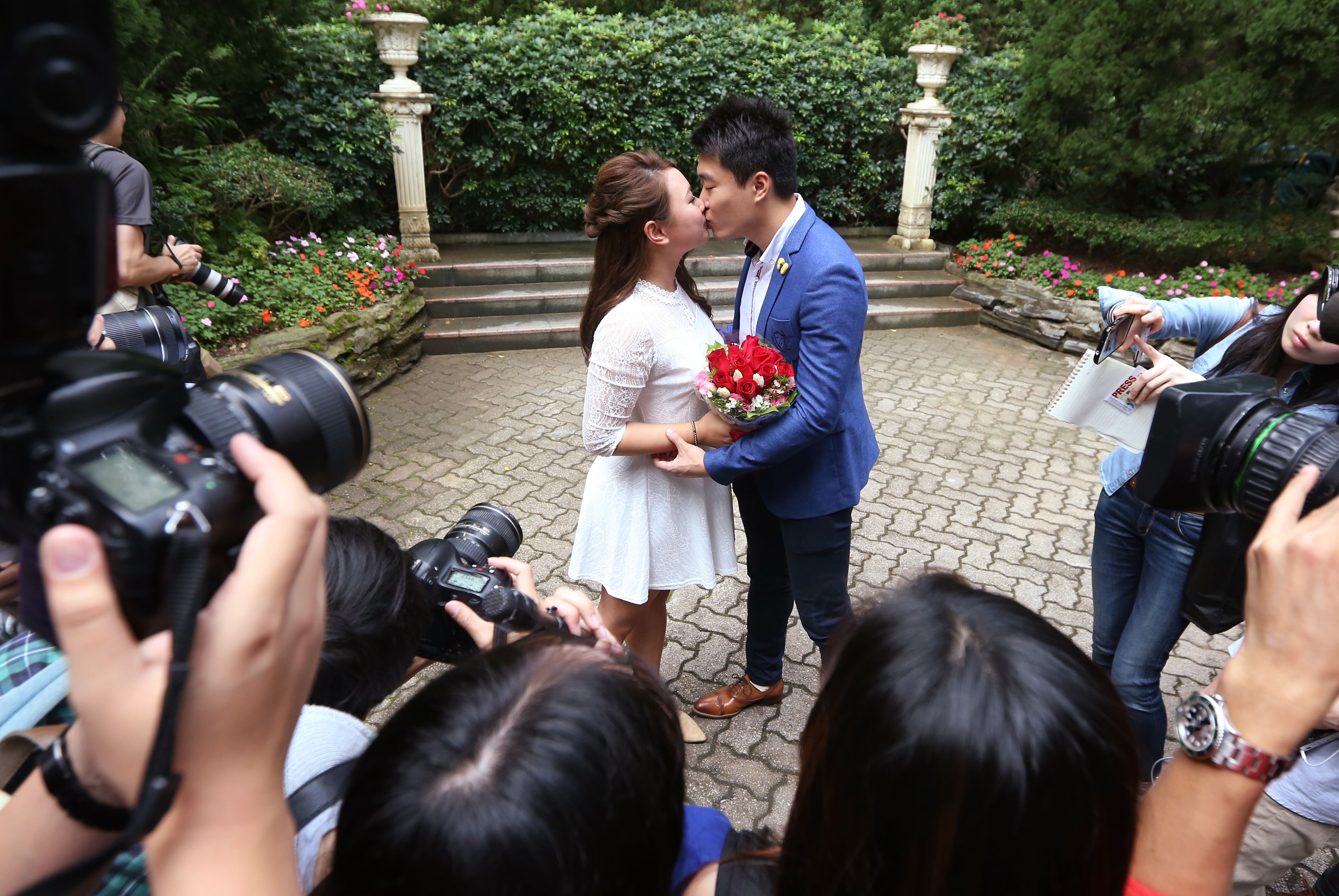 In this image, a man and a woman kiss in front of cameras in a park 