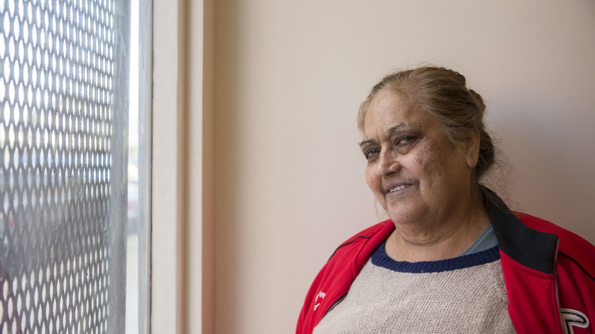 An older woman with gray hair tied back, wearing a cream sweater with blue stripes and a red jacket, stands beside a window with a metal mesh screen, smiling.