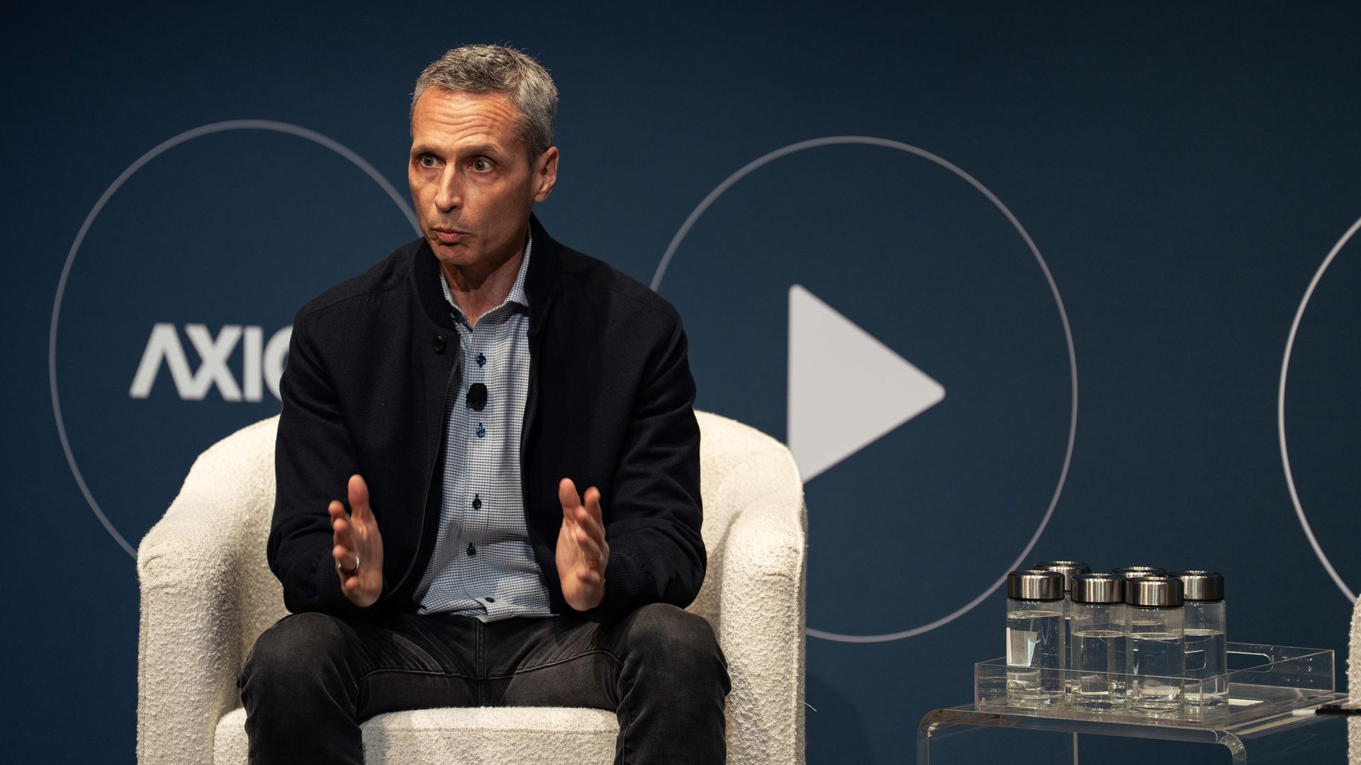ESPN chair Jimmy Pitaro in a black jacket and checkered shirt speaking while sitting in a white chair on stage, dark blue background at Media Trends Live.