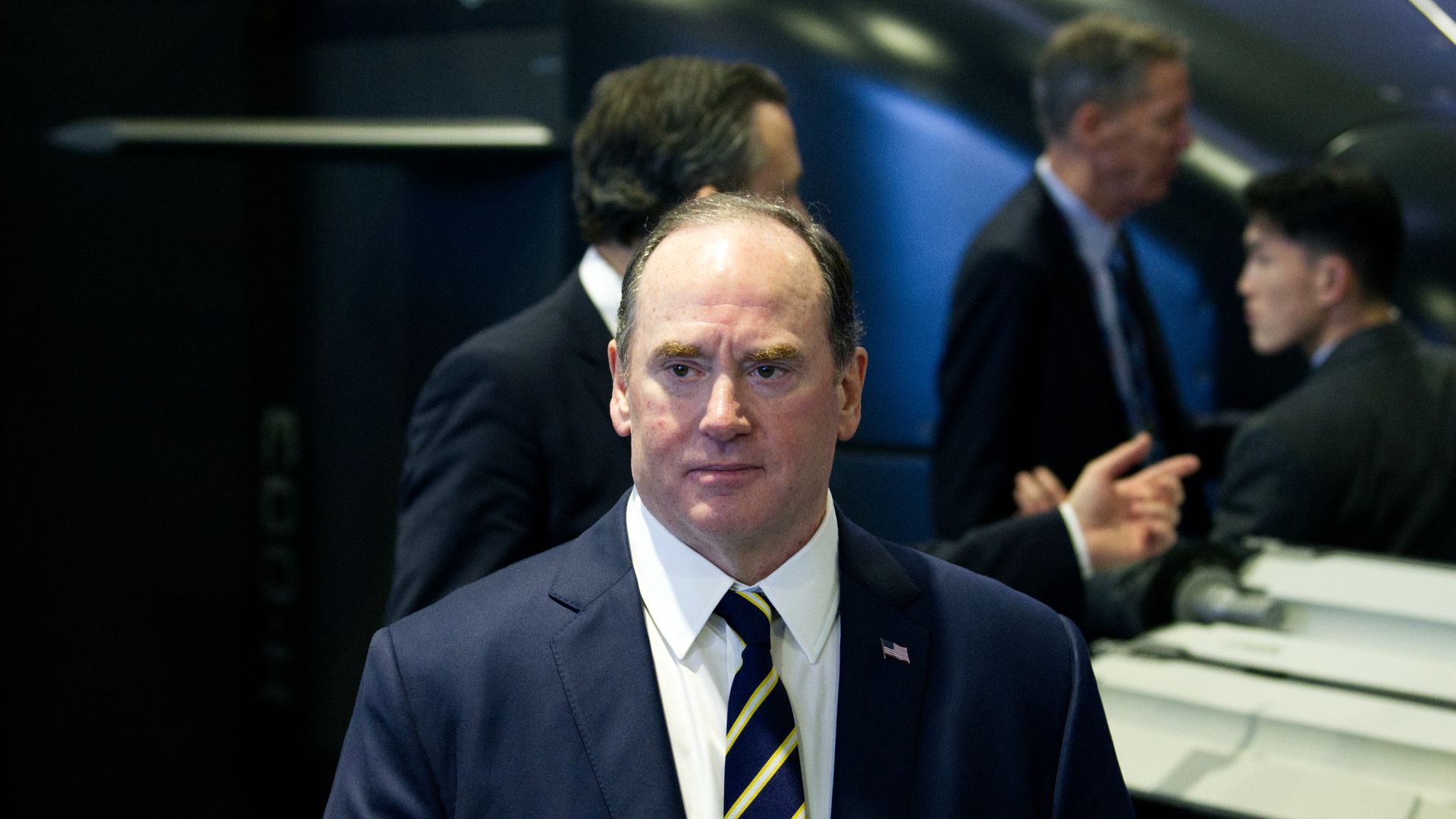 Man in a dark blue suit, white shirt, and striped tie with a small American flag pin, standing indoors with blurred men and large industrial equipment in the background.