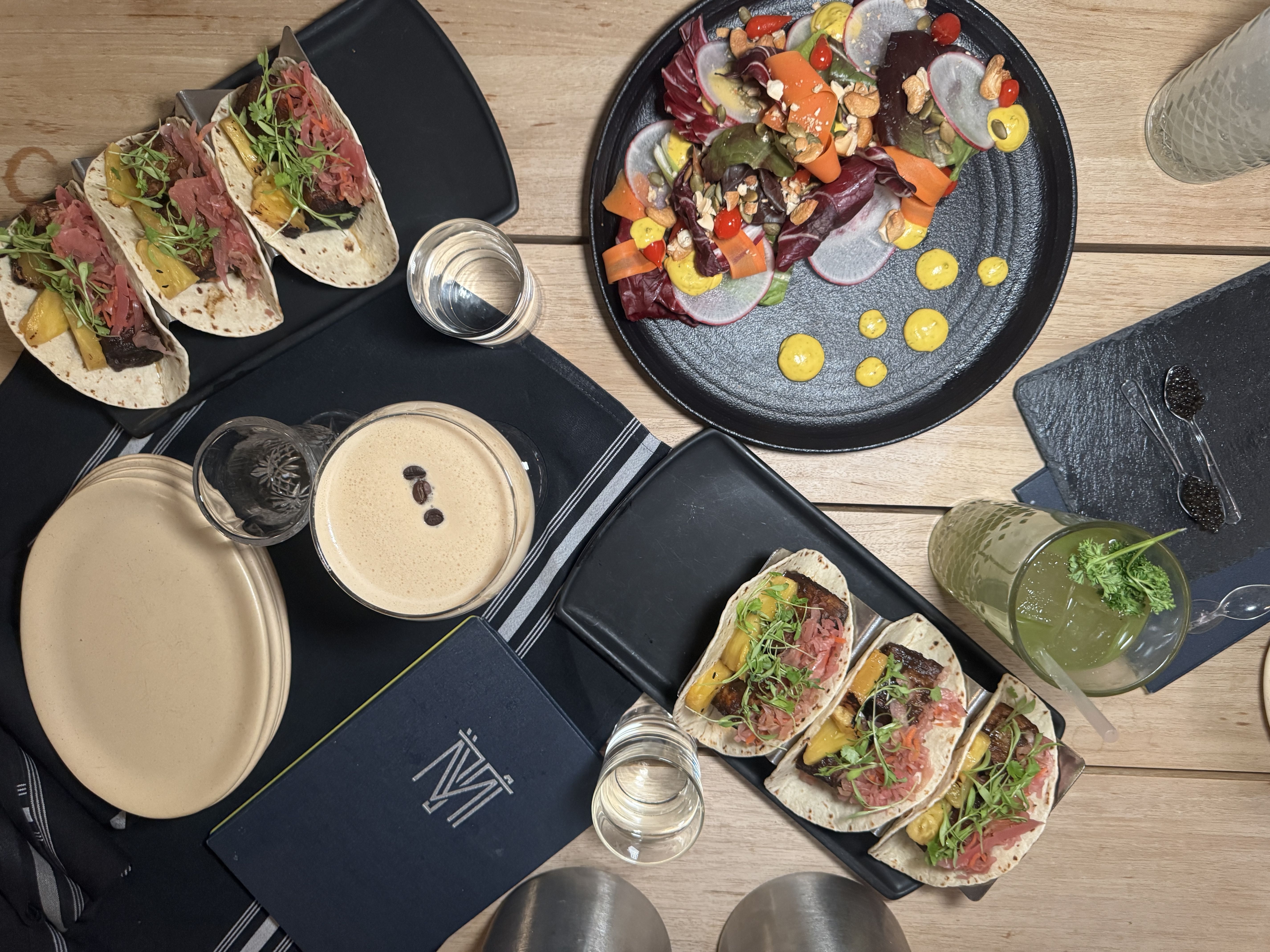 Overhead view of a wooden table with plates of tacos topped with greens and pineapple, a black plate with colorful salad and mustard dots, two drinks including a creamy one with coffee beans, and a menu.