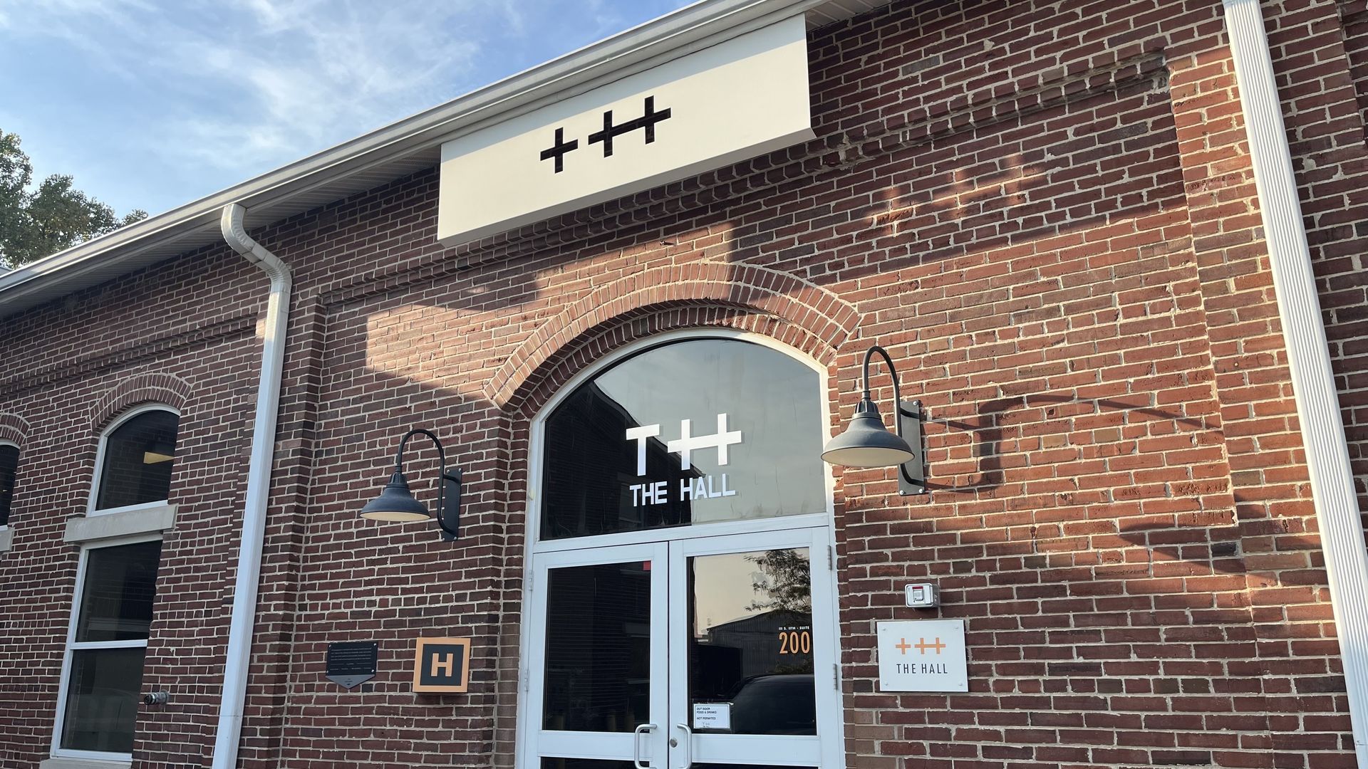 The front of The Hall, showing a brick building entrance with two black wall lamps, white double doors, and a sign above reading "+++"