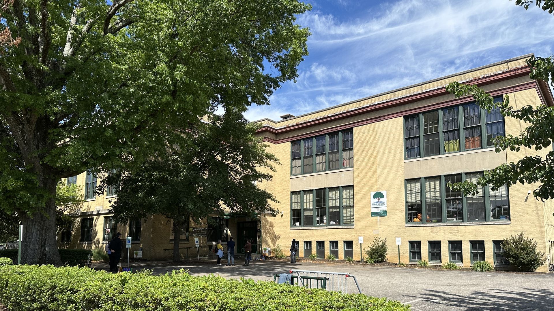 Beige brick building with large green-framed windows, partly hidden by a large leafy tree. Hedge in the foreground, a few people near the entrance, blue sky with wispy clouds.