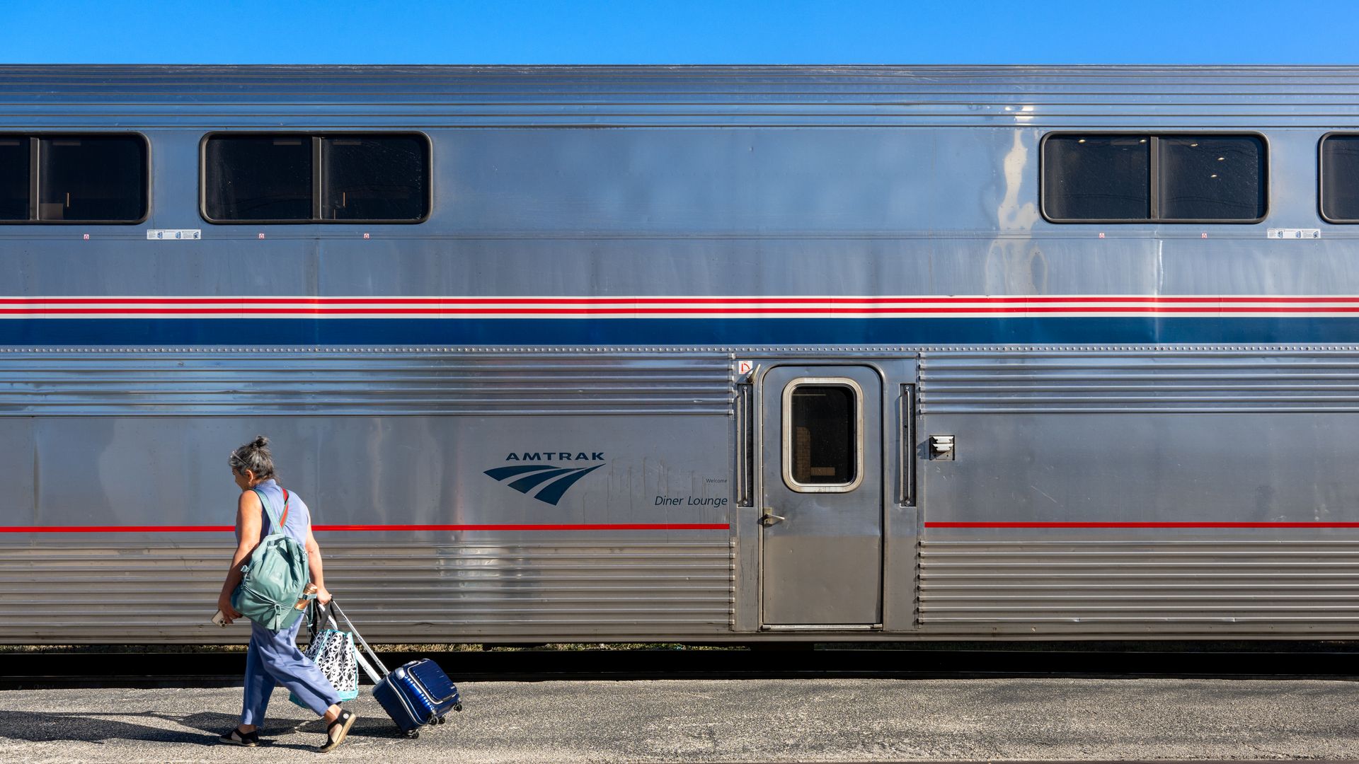 A person pulling a suitcase and carrying beside a silver Amtrak train car.
