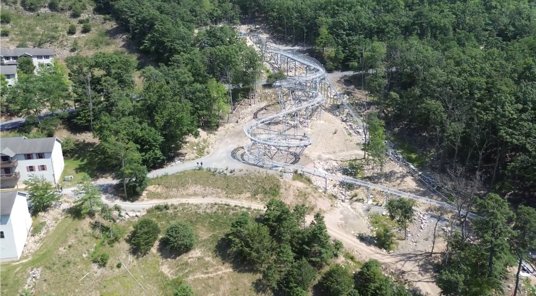 An aerial view of a gray steel roller coaster under construction in a forested hillside, with twisting tracks on tall supports and a nearby residential complex and dirt roads.