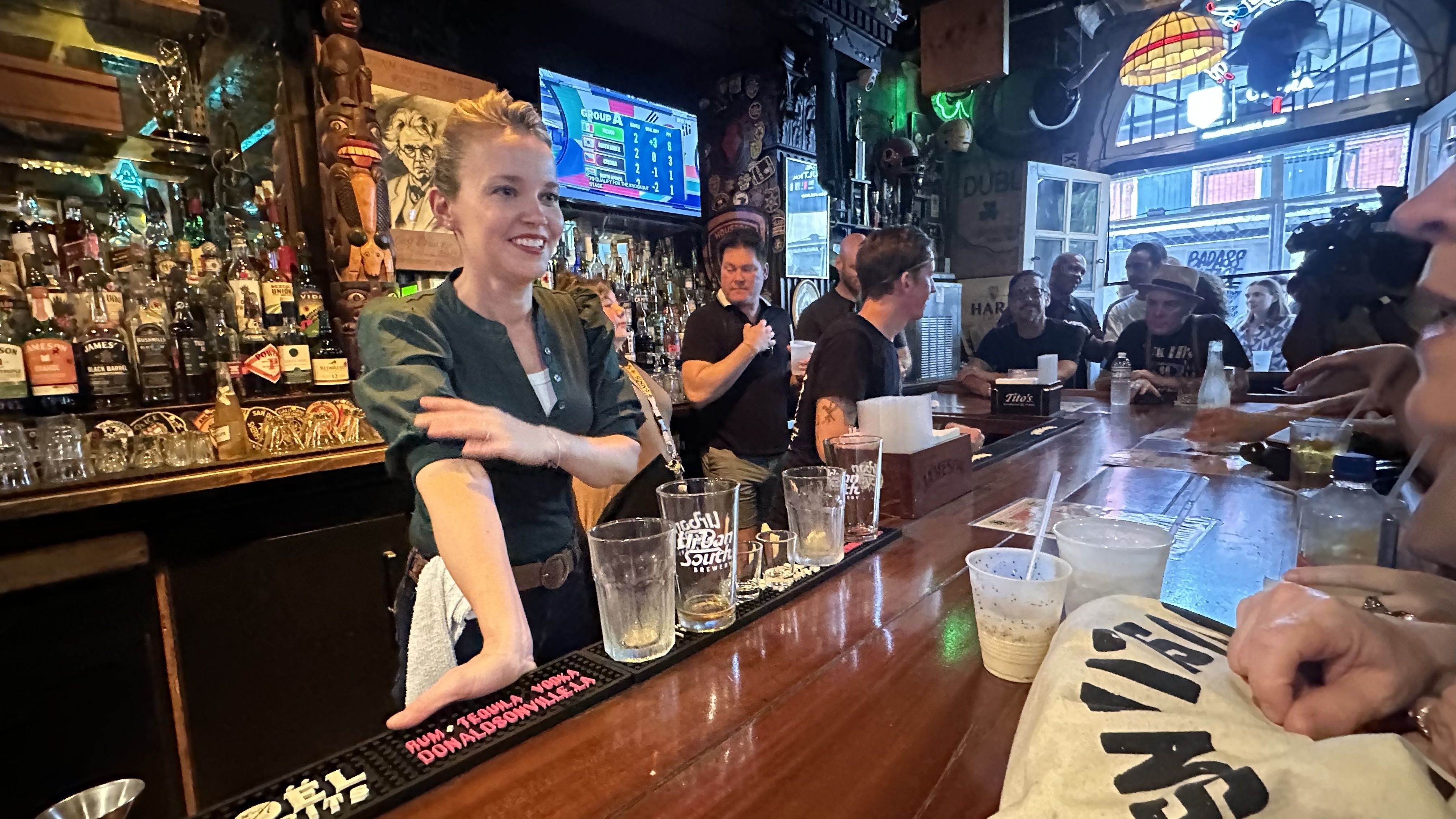 Smiling bartender in a green shirt serves at a crowded bar with many liquor bottles; a tiki statue stands behind, while patrons chat and a scoreboard TV glows in the back.