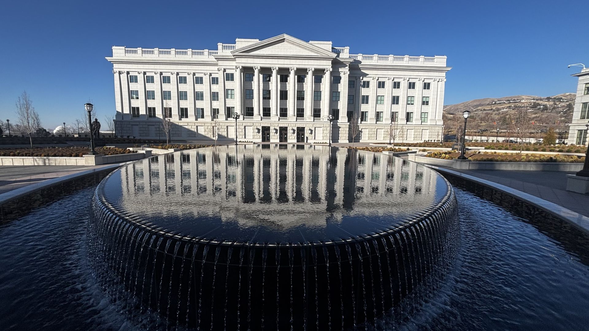 Large white neoclassical building reflected in a circular black infinity pool with water cascading over its edges, under a clear blue sky with distant hills and landscaping.
