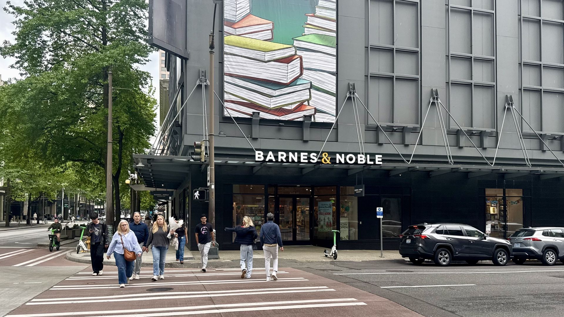 Facade of a Barnes & Noble store with a tall banner showing stacked books and the words "Begin Here"; pedestrians cross a wide crosswalk beside trees and a gray city building.