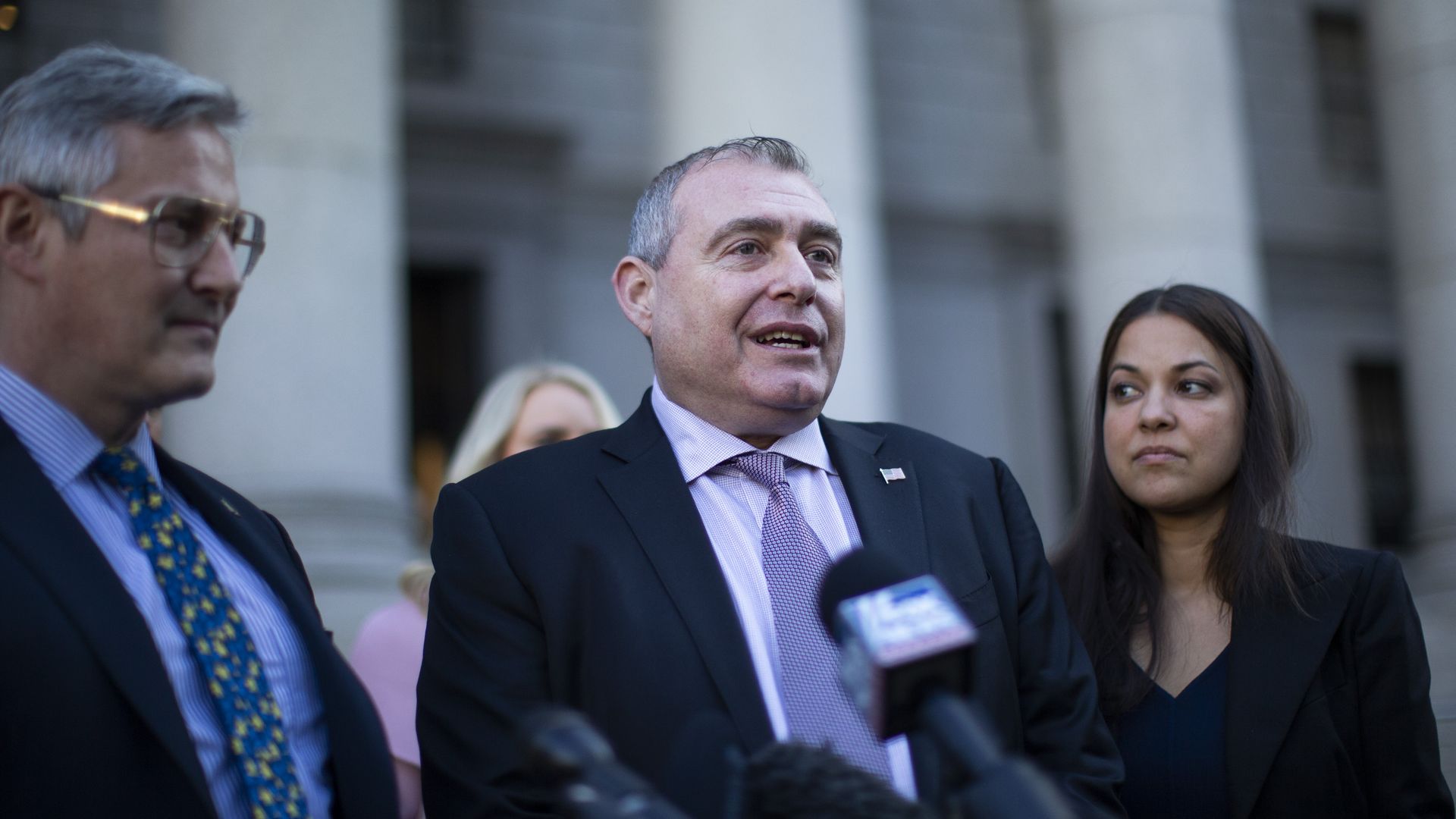 Lev Parnas, wearing a dark blue suit, light purple shirt and purple tie, speaks into microphones while flanked by his lawyers in front of a white marble courthouse.