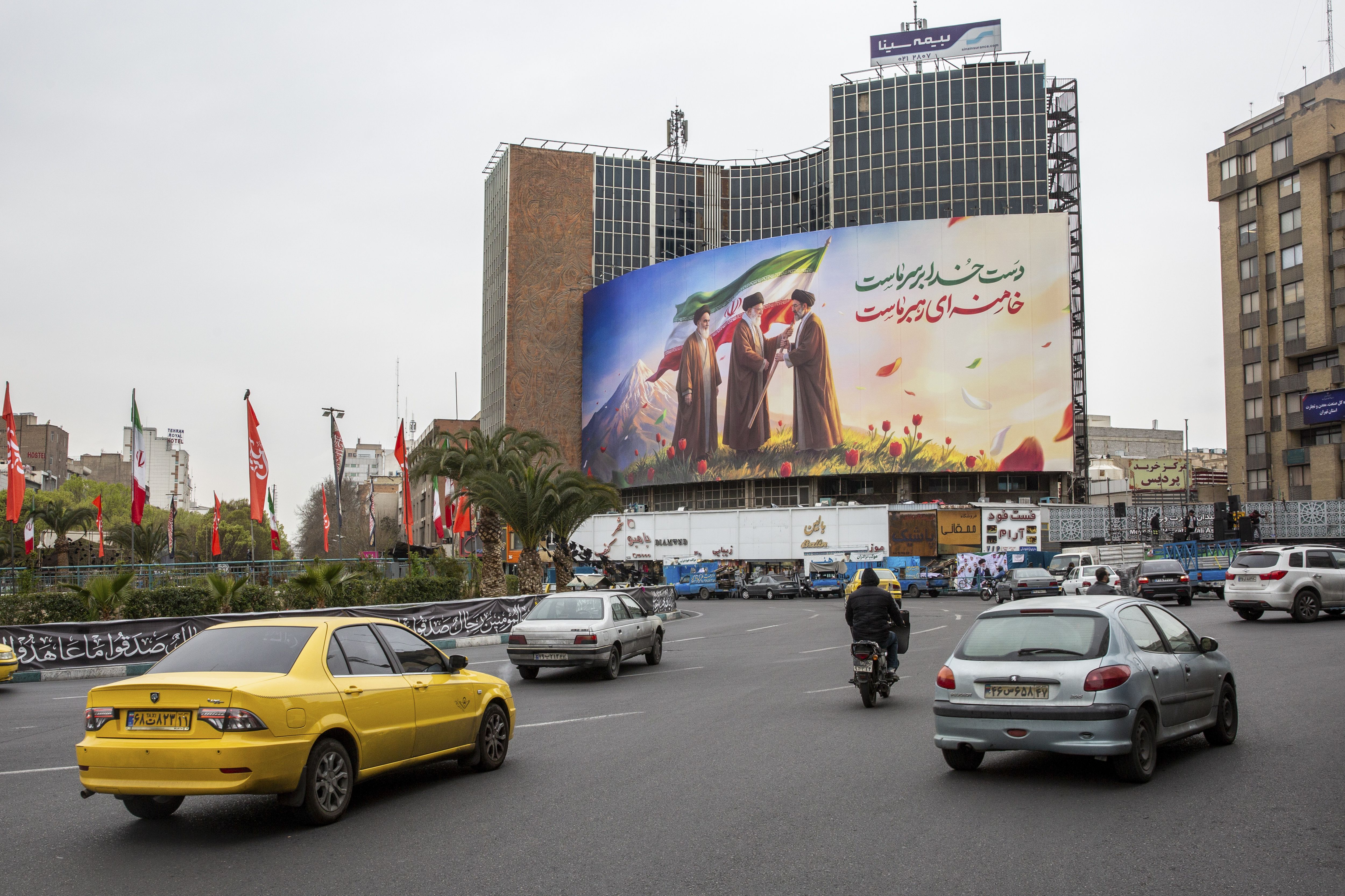 A new billboard in Valiasr Square in Tehran shows the late Ruhollah Khomeini (left) looking on as Ayatollah Ali Khamenei (center), killed on Day 1 of the war, passes an Iranian flag to his son, Mojtaba Khamenei, the new supreme leader. Photo: Arash Khamooshi for The New York Times