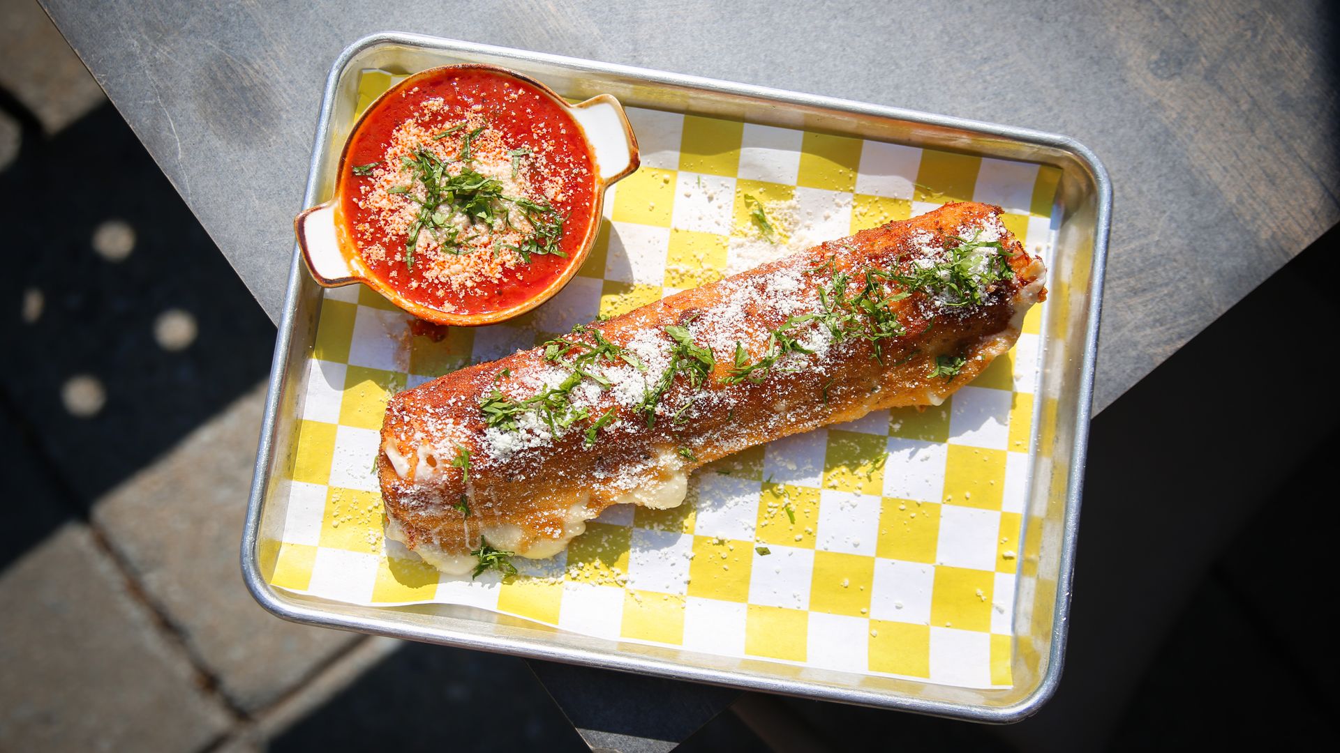 Metal tray with yellow checkered parchment holds a long fried mozzarella stick sprinkled with parsley and cheese, next to a red tomato soup in a small orange bowl.