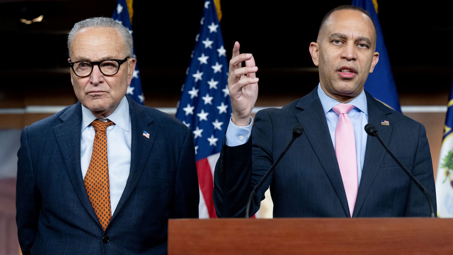 Two men in suits with American flag pins stand at a podium with microphones. The man on the left has glasses and an orange tie, the man on the right speaks with a pink tie and raised hand.