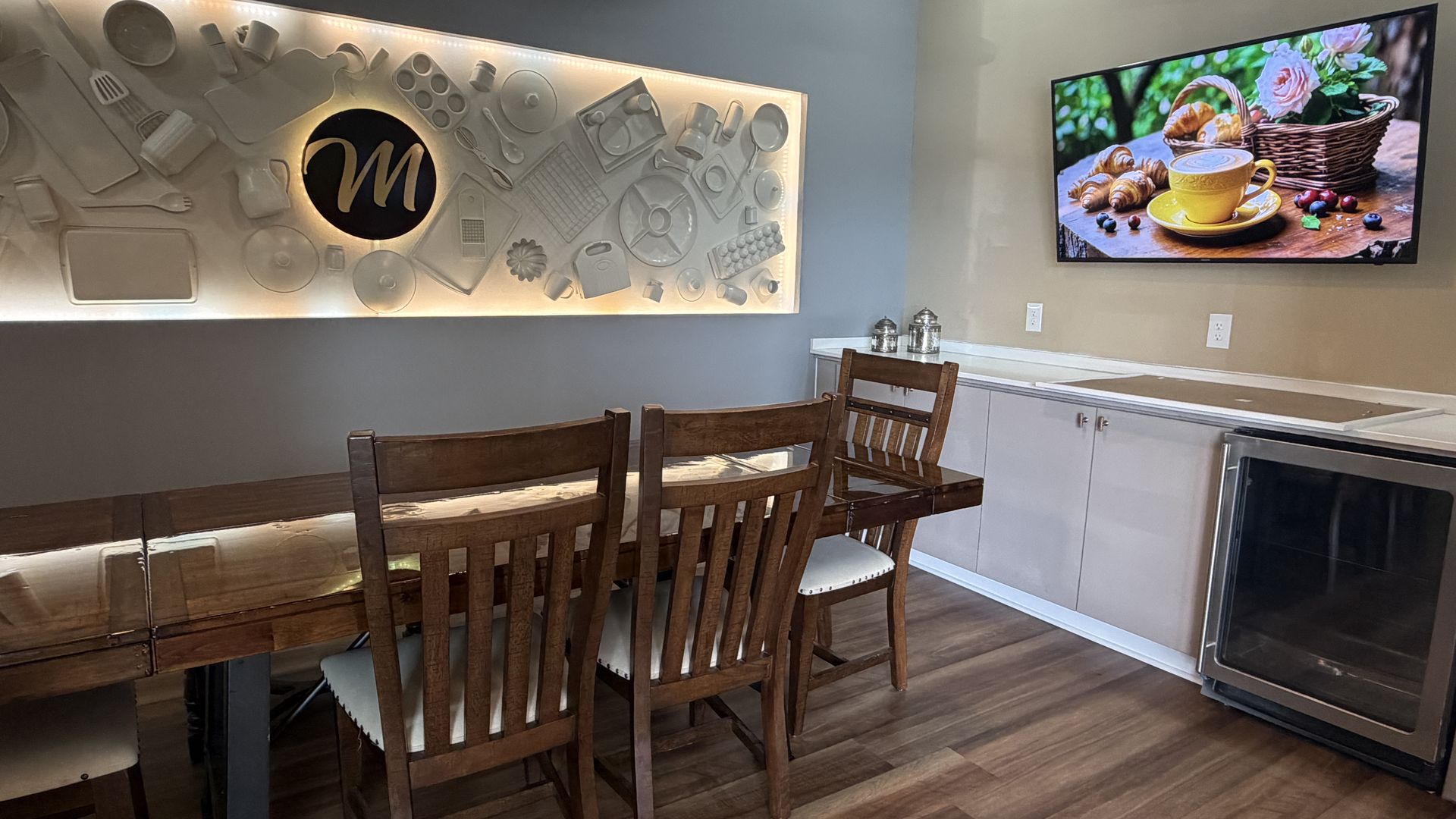Cafe interior with a long wooden table and chairs. A backlit mural of kitchen tools and an M logo on the wall. A TV shows coffee and croissants; beige walls, wood floor, and stainless appliances.
