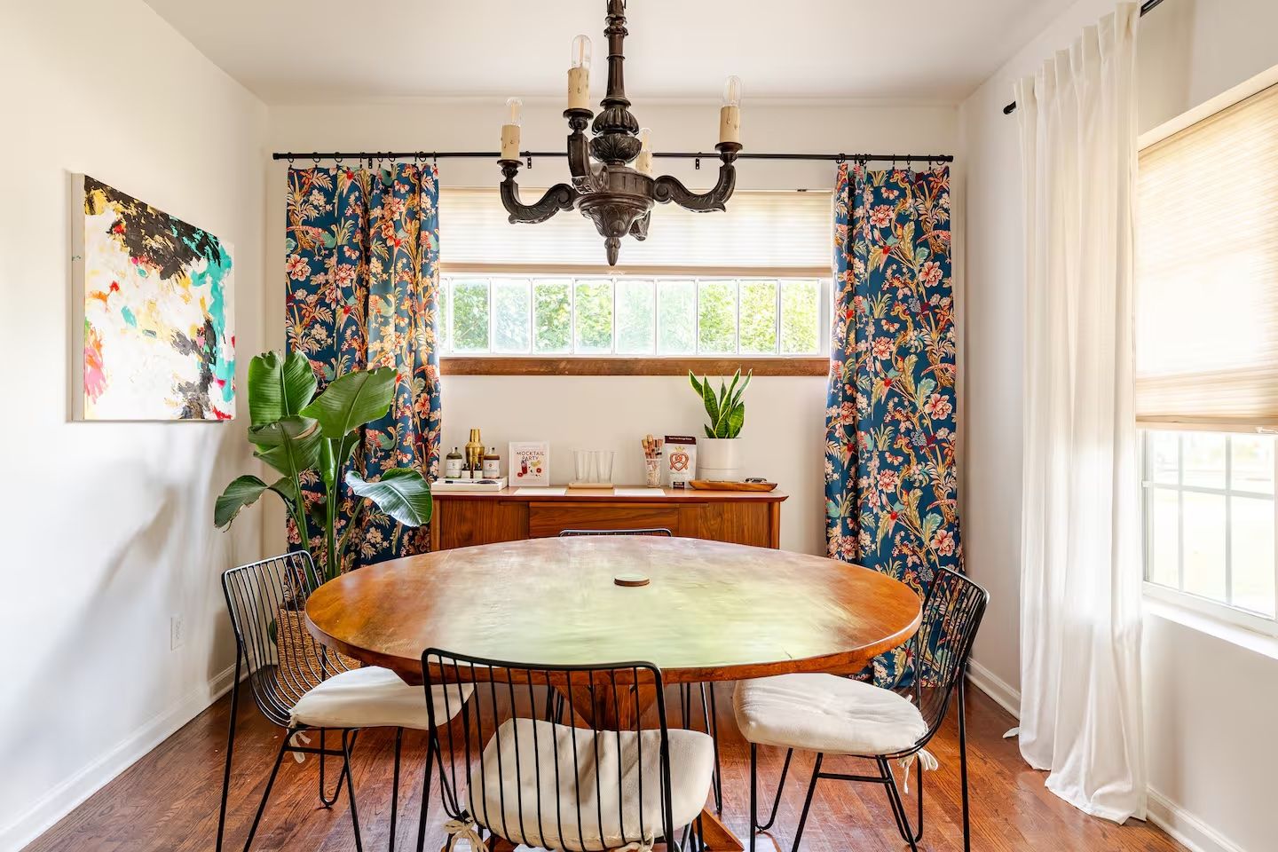 Bright dining room with round wooden table, four black metal chairs with white cushions, large window with floral blue curtains, white walls, leafy green plant, and black chandelier.