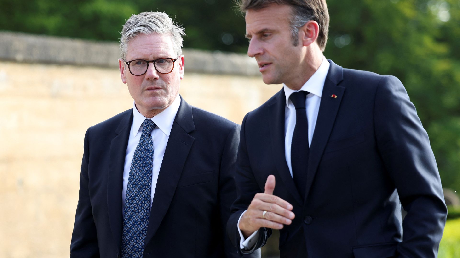 Two men in dark suits, one with gray hair and glasses, the other with brown hair, walk outdoors near a stone wall and green trees, engaged in conversation.