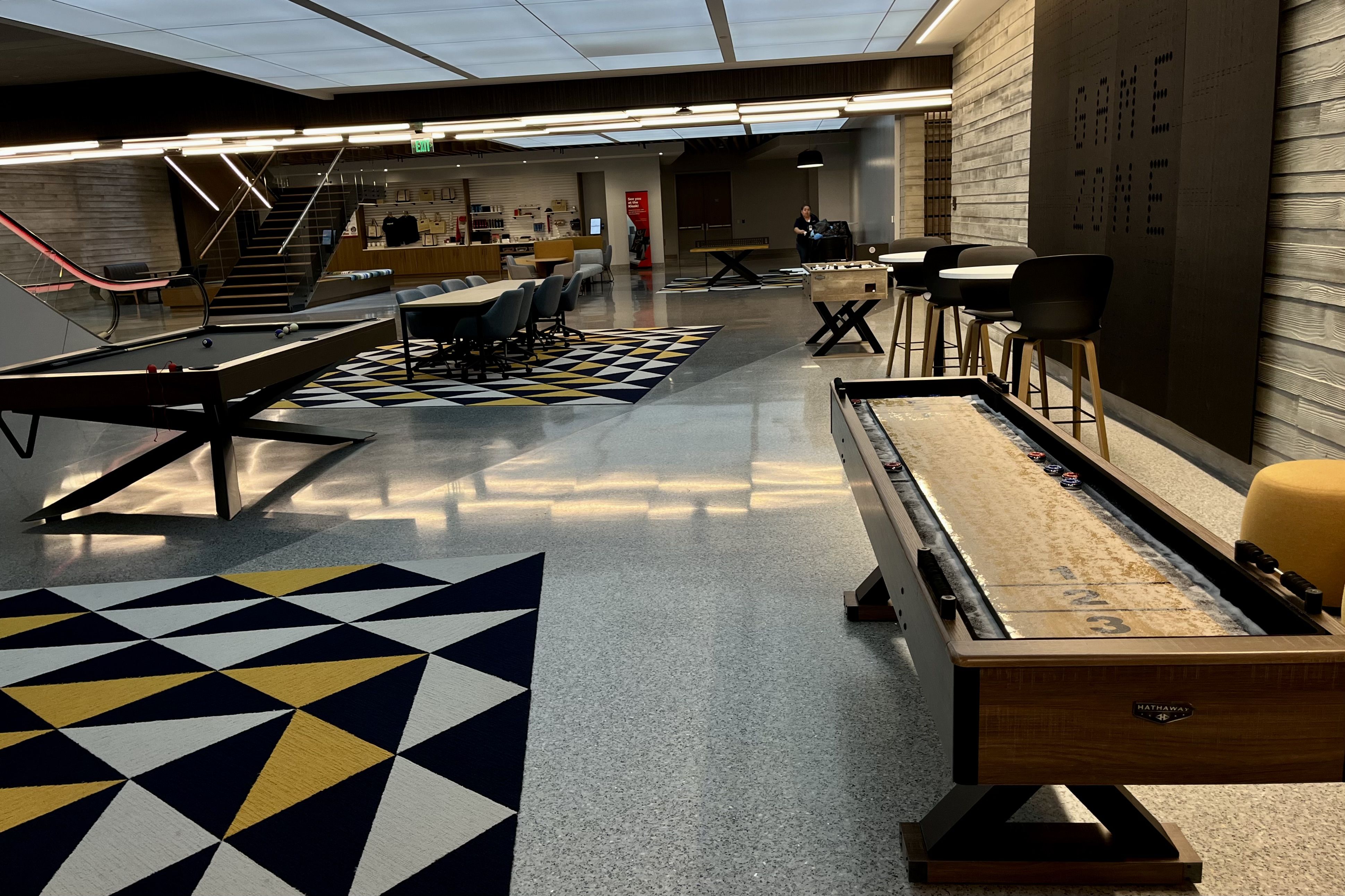 Modern game lounge with a pool table, shuffleboard table, foosball, high stools, geometric patterned rugs in navy, white, and yellow, and a person cleaning in the background.