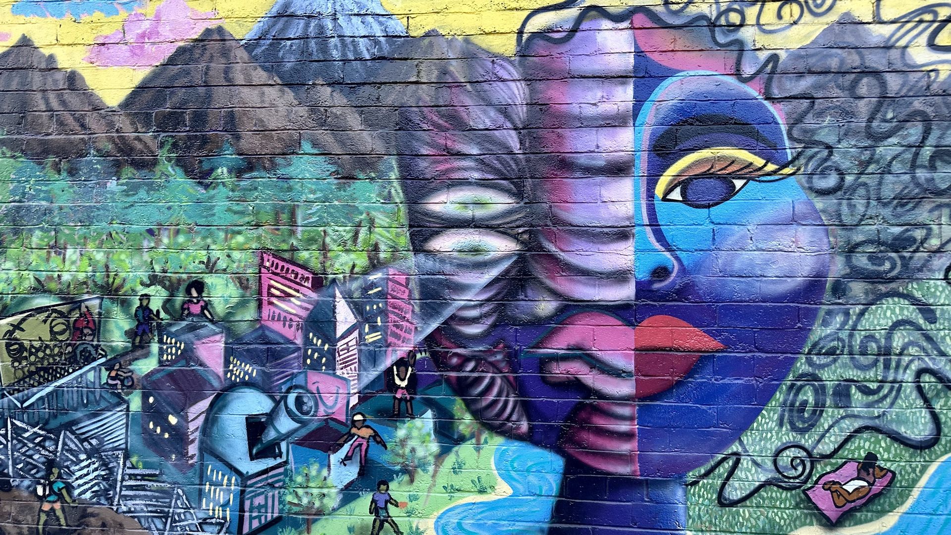 A mural featuring a yellow sky, pink clouds, mountains in the background and a purple and blue face in the foreground, with a city in between.