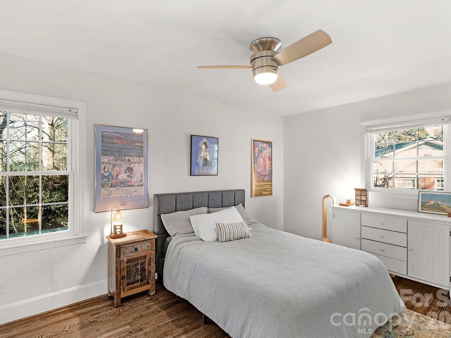 Bright bedroom with white walls, wood floor, gray upholstered bed with gray bedding, wooden nightstand, dresser under window, ceiling fan, and art posters on the walls.