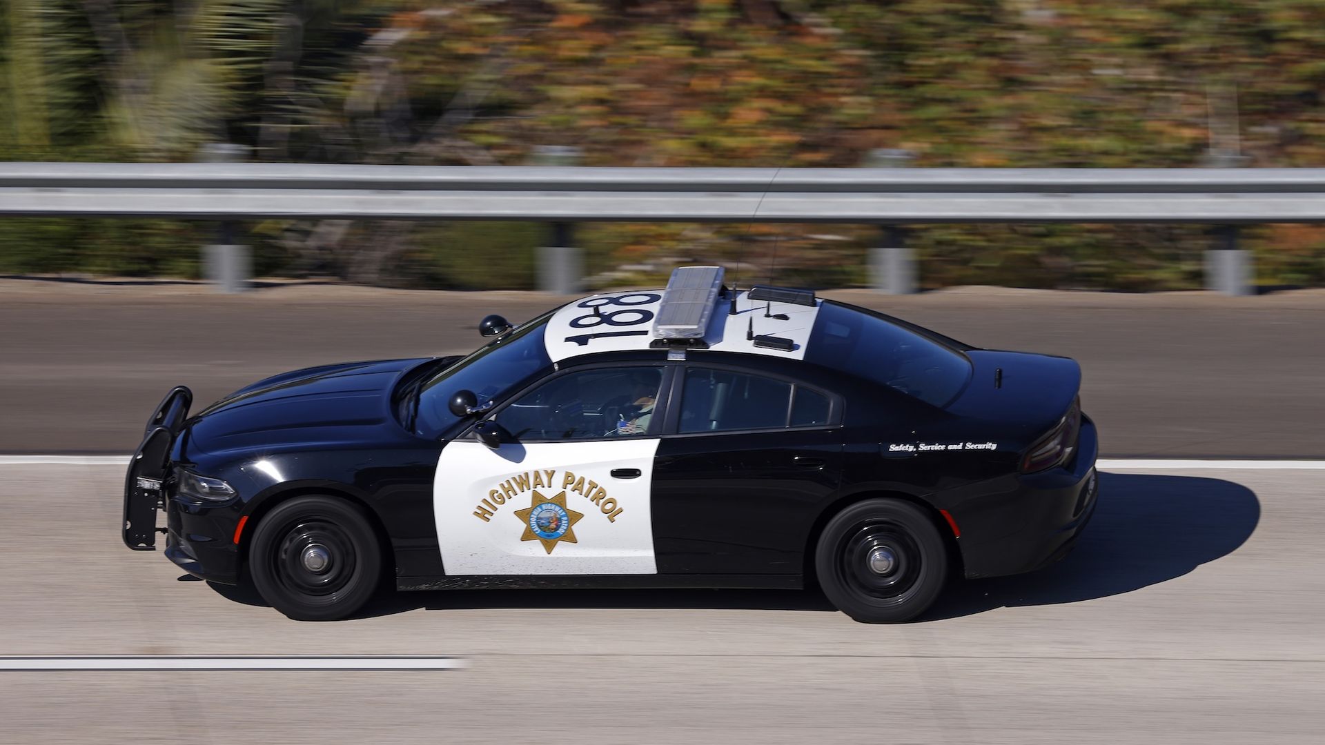 A California Highway Patrol car driving on a highway past a guardrail with greenery in the background.