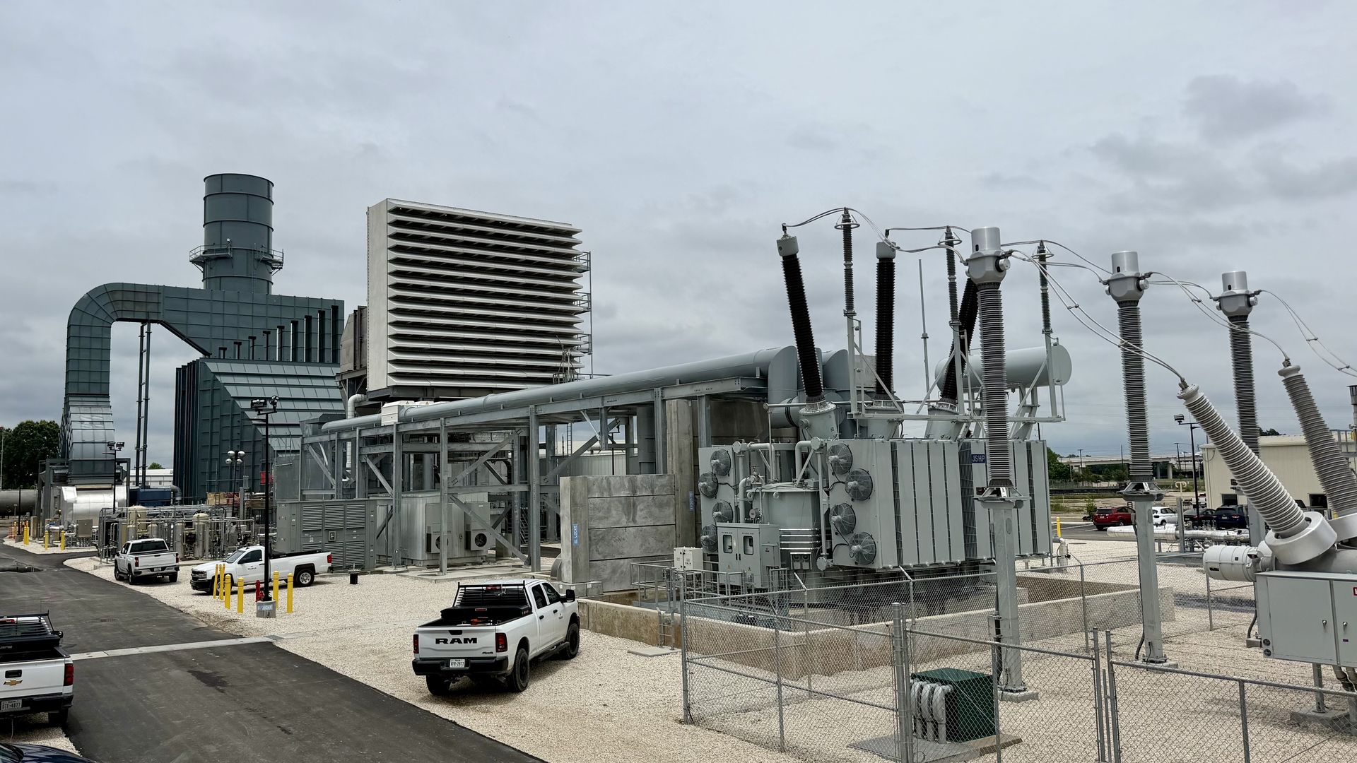Power plant with electrical equipment: a cooling tower, large vent louvers, transformers, insulators, and metal piping. A chain-link fence, vehicles, and a cloudy sky.