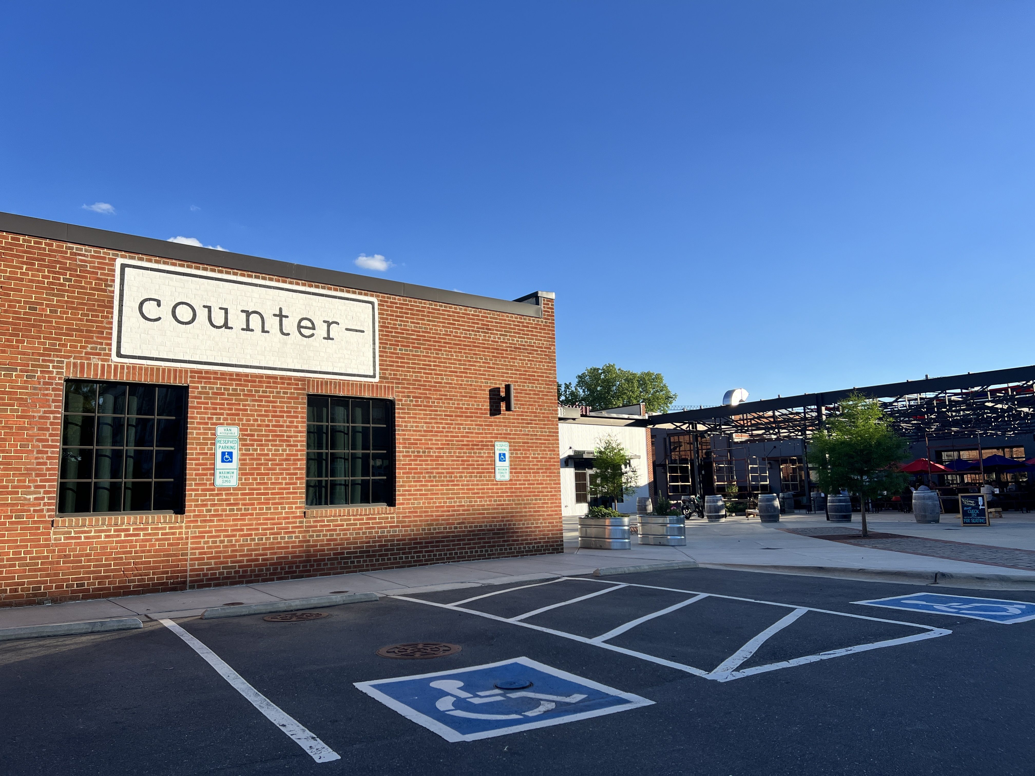Red brick building with a white sign reading "counter-" above two black-framed windows, adjacent to a patio area with trees and seating, under a clear blue sky, and handicap parking spots in front.