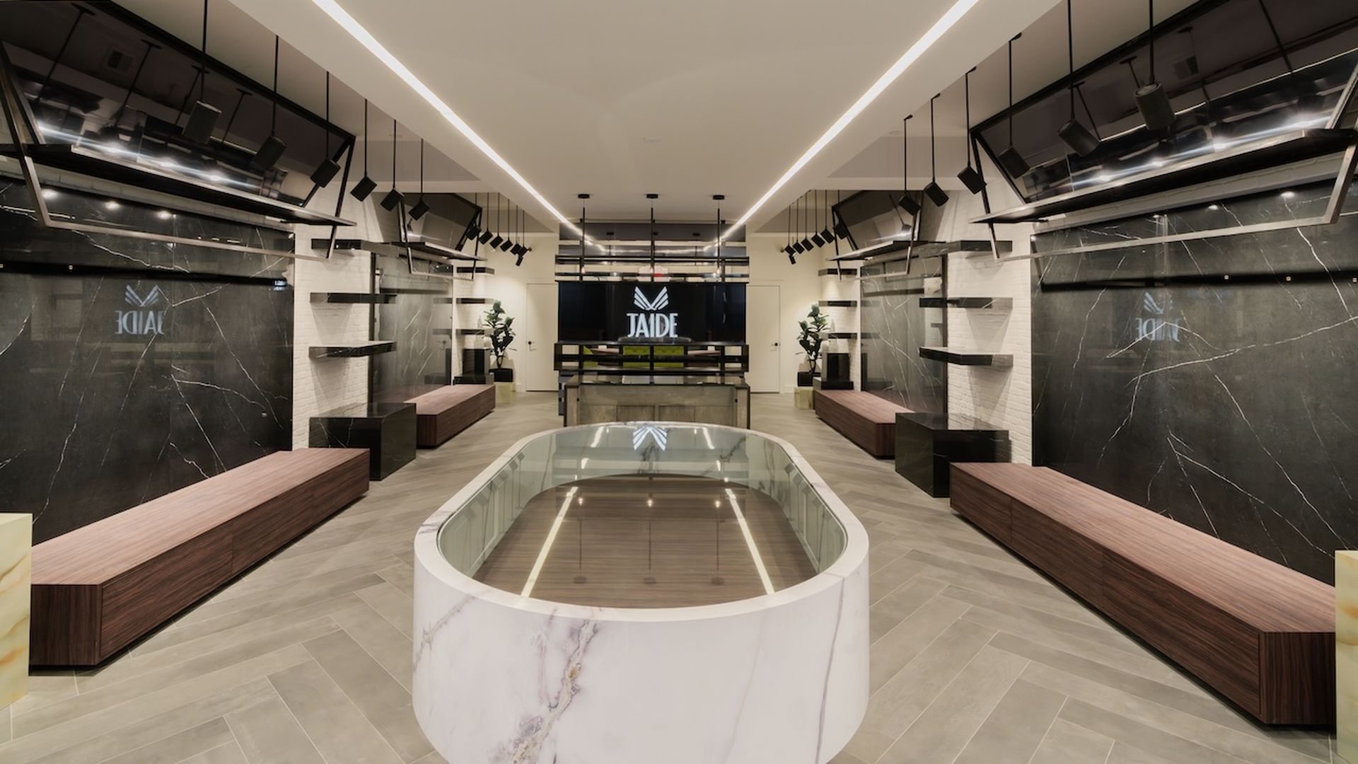 Modern, symmetrical interior with marble-topped central table, wood benches, black marble walls with shelves, large flat screens, and ceiling lights, labeled "JAIDE" at the back.