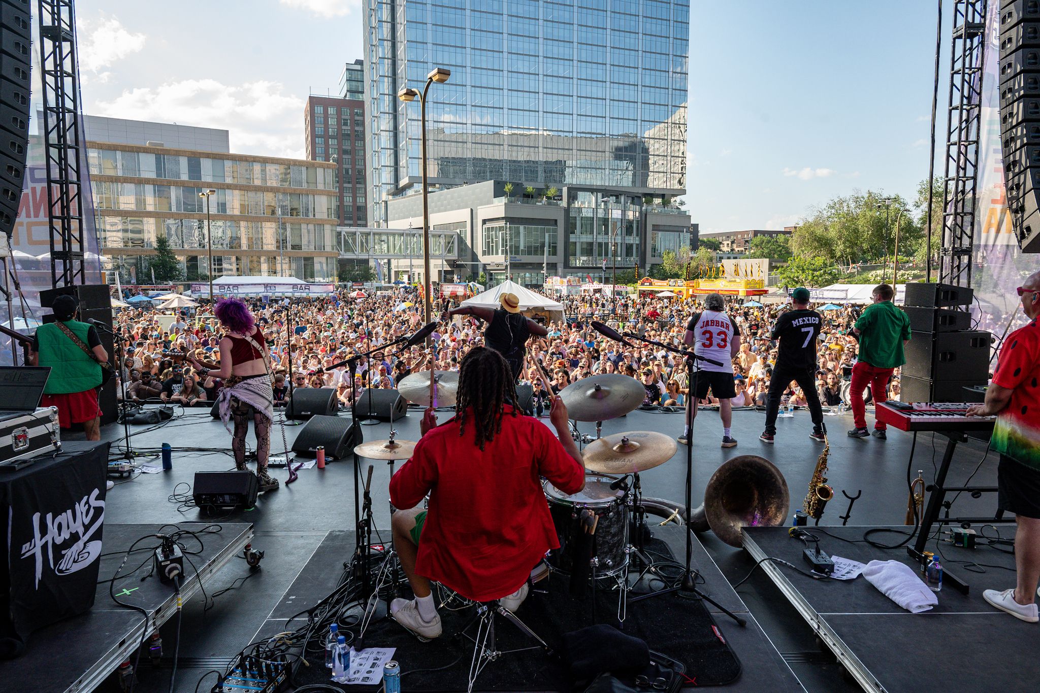Backstage view of a large outdoor concert in a city plaza; musicians perform on stage as a dense crowd fills the square, with tall glass buildings and blue sky in the background.