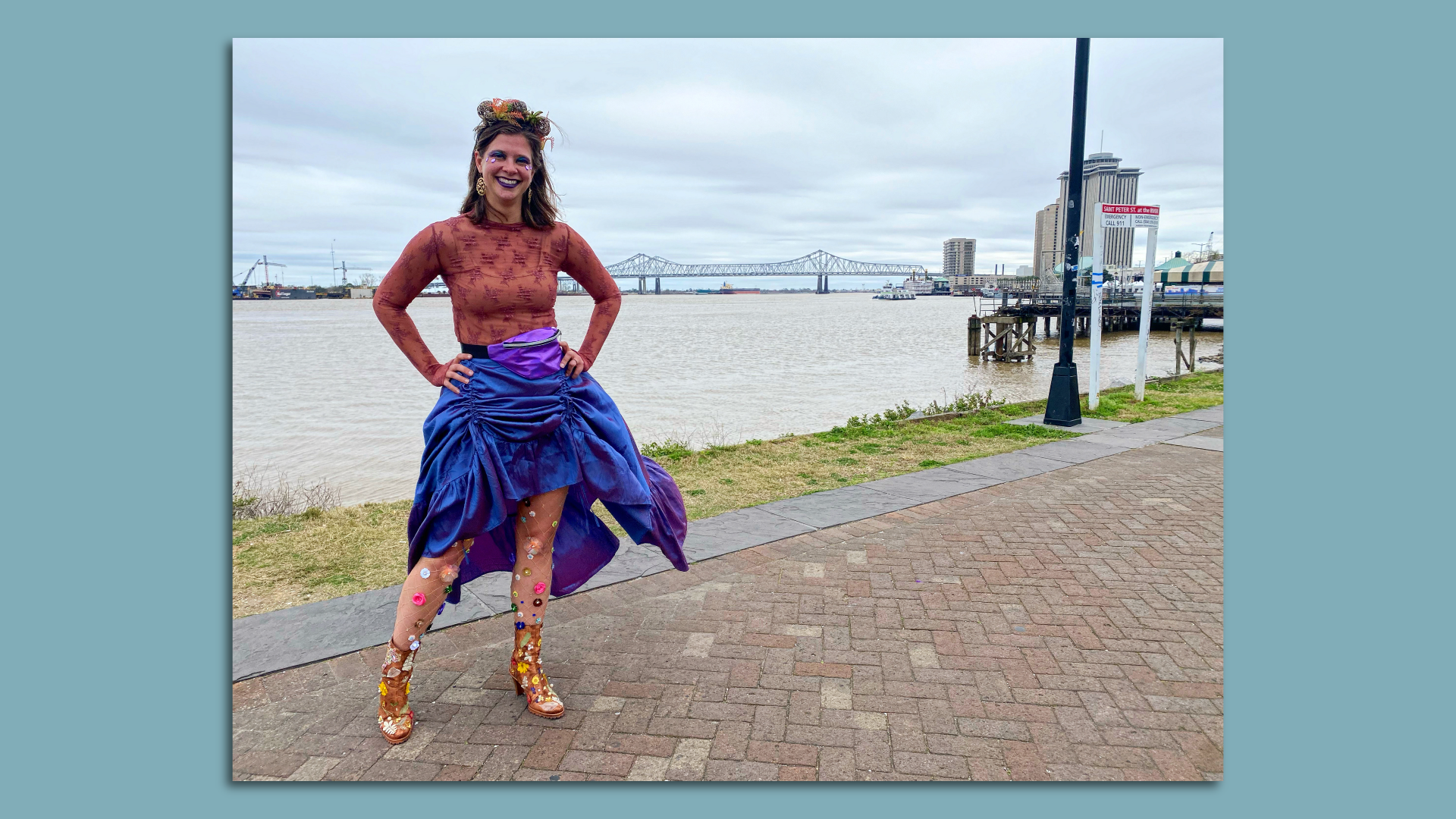 A woman stands on the New Orleans Moon Walk, a pathway next to the Mississippi River, with the Crescent City Connection bridge in the background. She wears a floral costume, with boots covered in faux flowers.