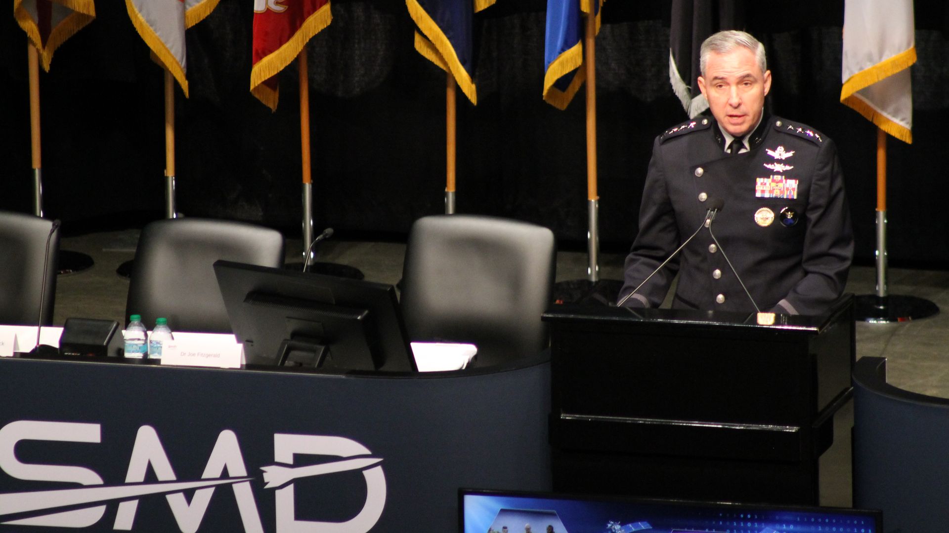 Senior military officer in dress uniform speaks at a conference podium; several flags on poles line the dark stage behind him, with conference signage in the foreground.