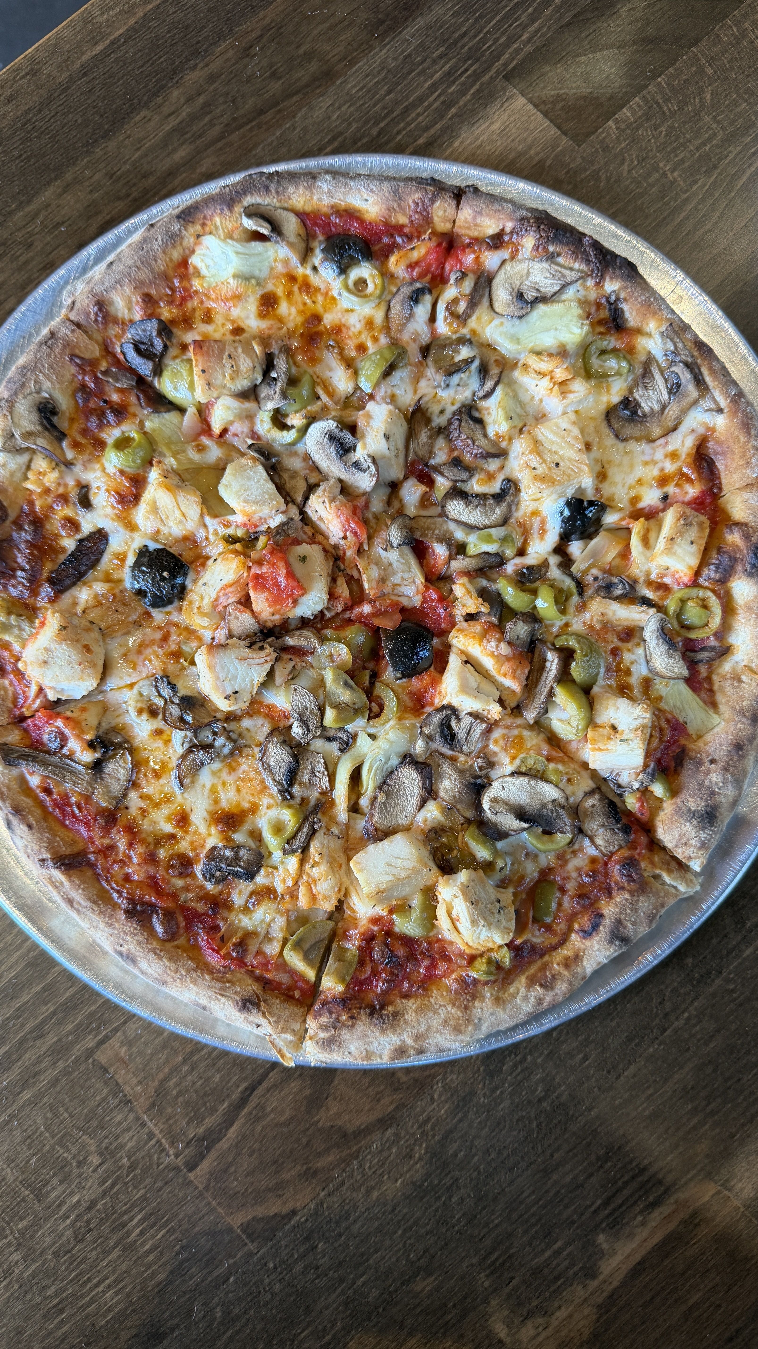 Large round pizza on a metal tray, with melted cheese and tomato sauce, topped with chicken, mushrooms, green olives, pineapple, and peppers.