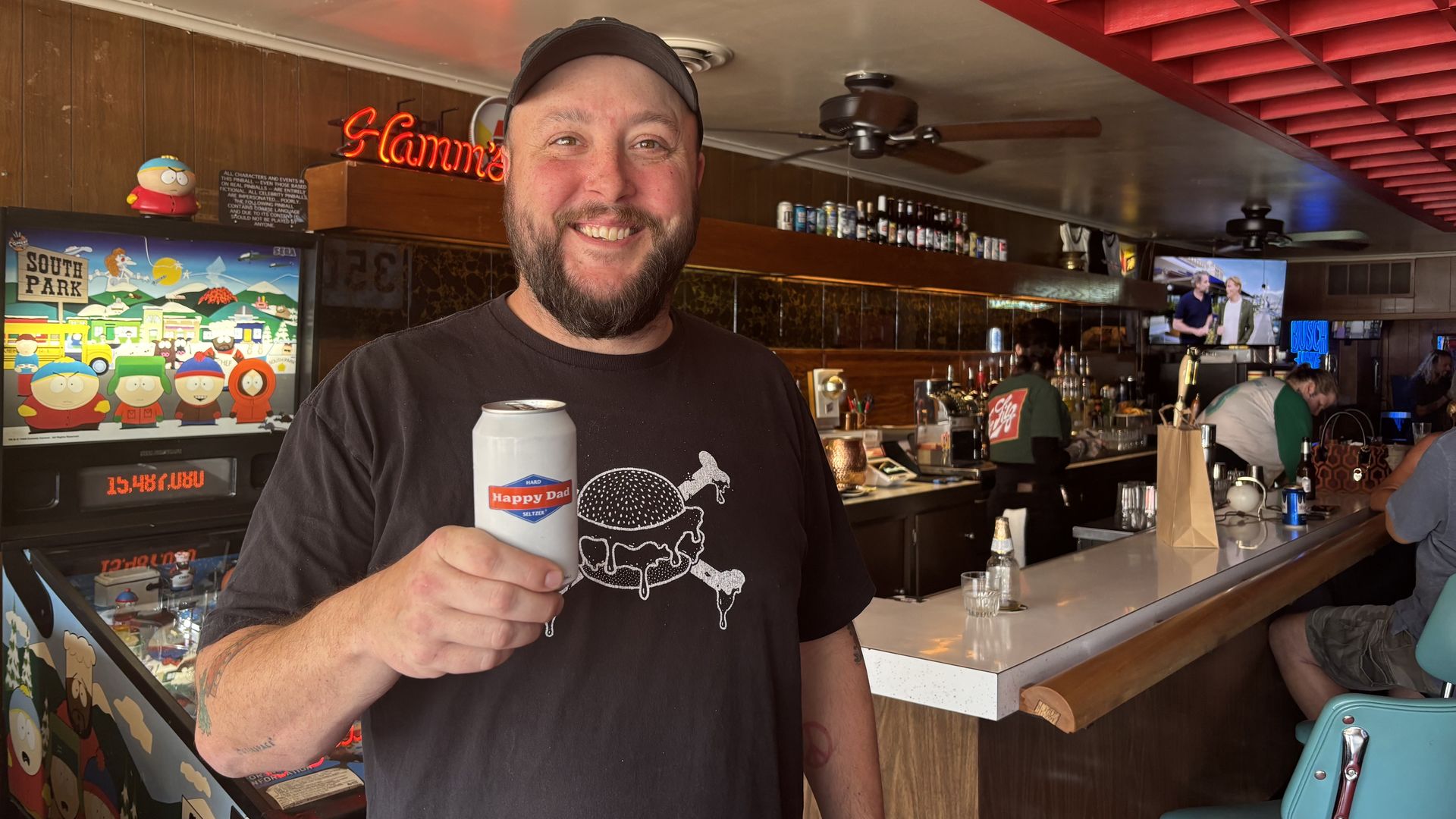 Cory Wendel smiling in a black cap and t-shirt holds a can labeled "Happy Dad" inside a bar with a red ceiling grid, a South Park pinball machine, and patrons at the counter.