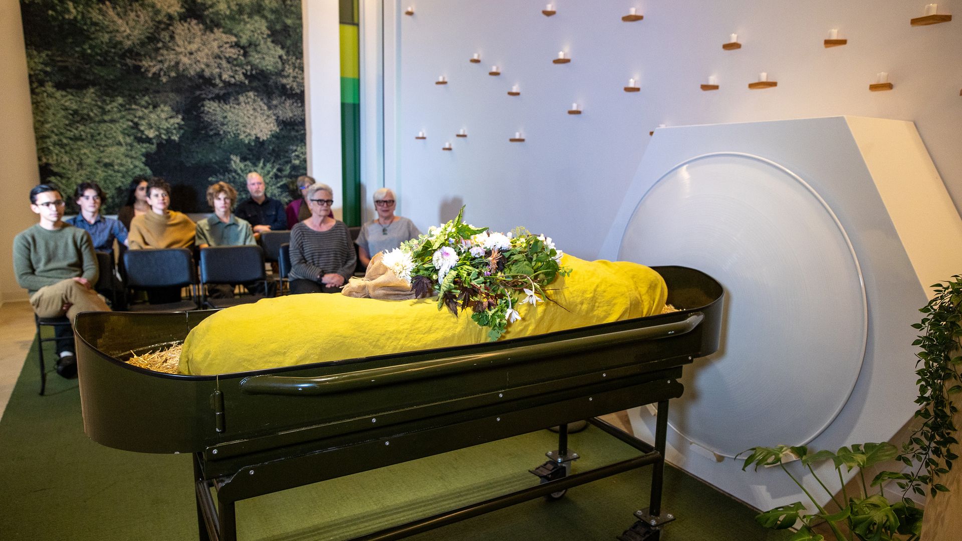 A mannequin wrapped in a yellow shroud sits with flowers in a funeral-like setting, in front of a large circle where the body will be placed for human composting. People watch in seats as at a funeral. 