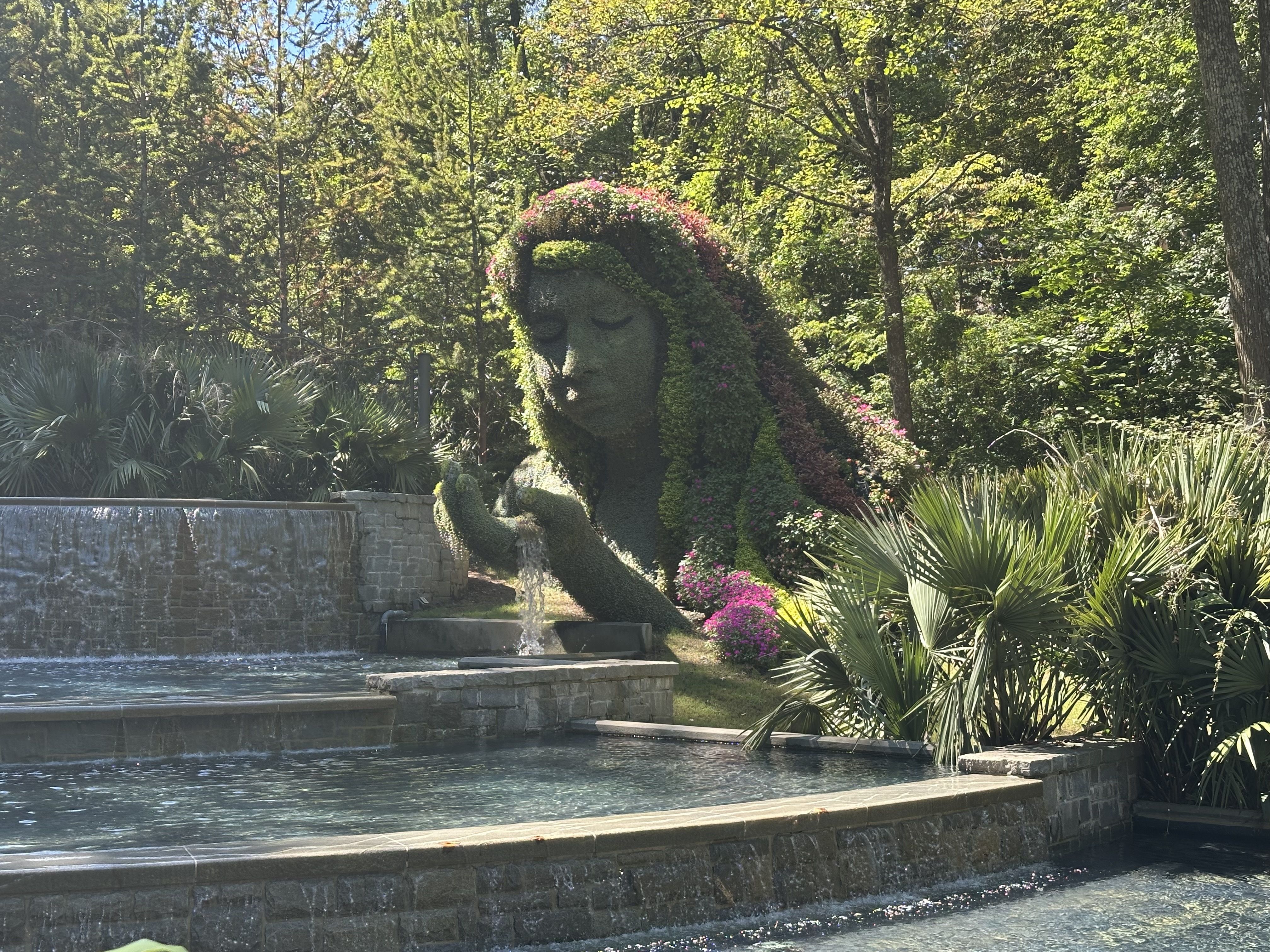 An ivy-covered sculpture of a serene, closed-eyed head rises beside a tiered stone fountain, pink flowers along its crown, with lush trees and spiky palms in the background.