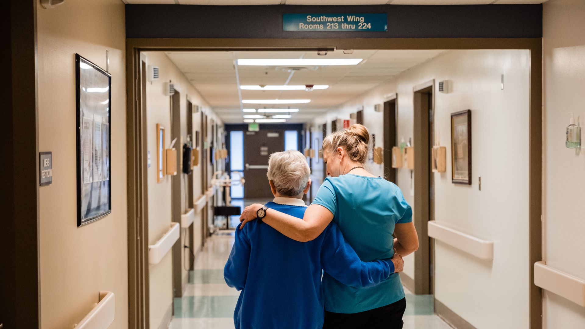 A nurse walks an elderly woman down an assisted living hallway.