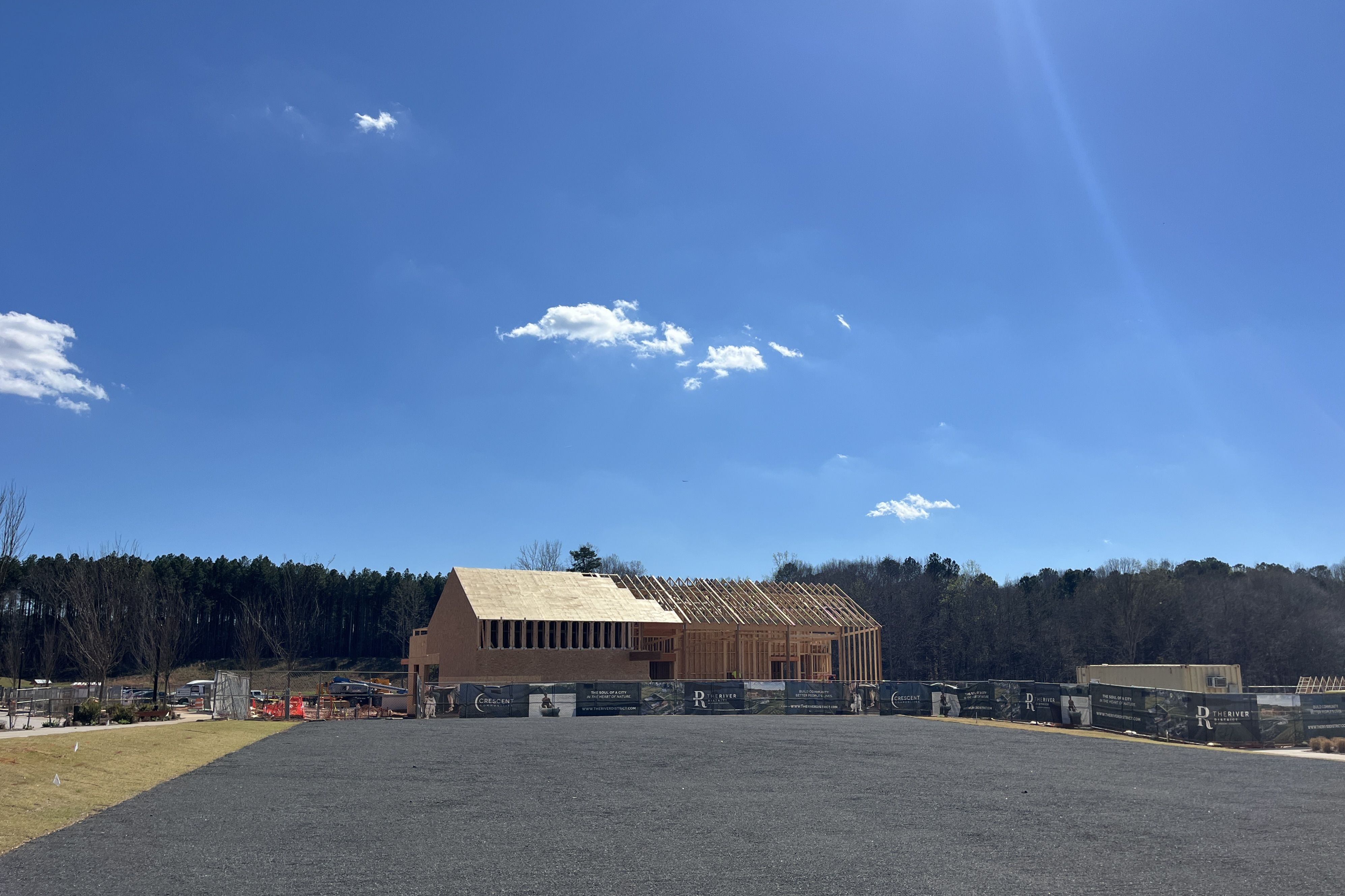 A sunny blue sky over a construction site featuring a wooden-framed house in progress on a gravel lot, fenced with banners; a tree line forms the distant background.