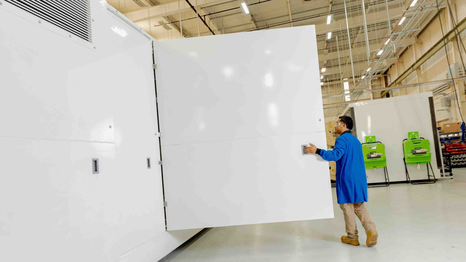 A worker in a blue lab coat and brown pants opens a large white door to a white box in a pristine factory floor.