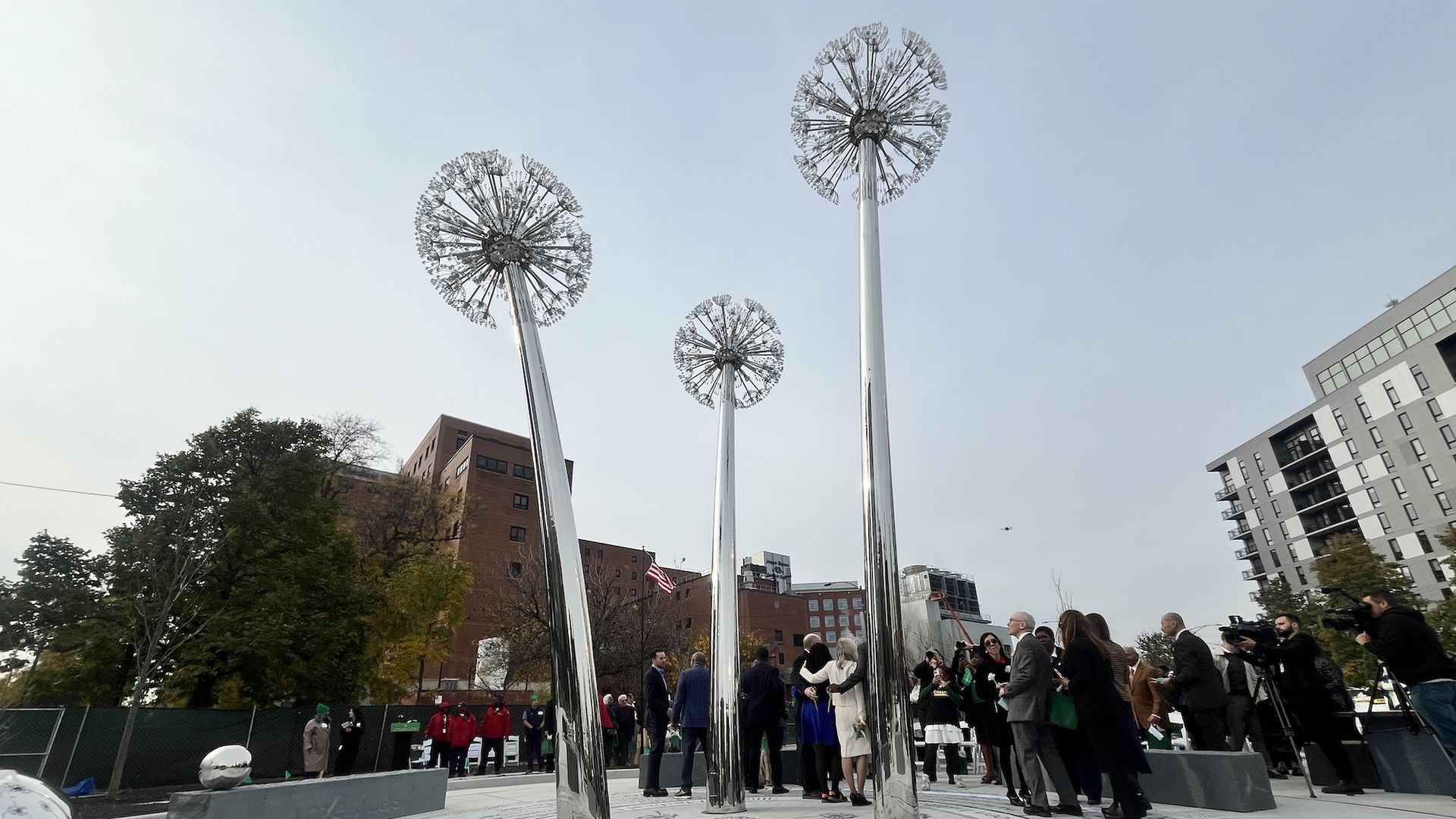 Polished metal poles with round, dandelion-like toppers rise from a circular granite memorial etched with text; a crowd of people in formal attire gathers while an American flag waves near buildings.