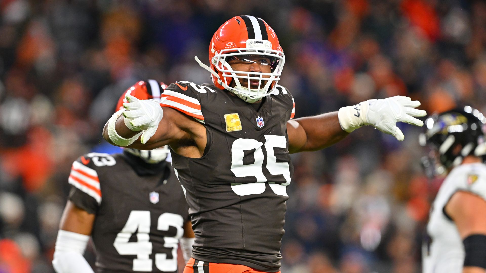 Cleveland Browns football player in an orange helmet and brown jersey with white gloves, arms outstretched in celebration on the field, blurred crowd in the background.