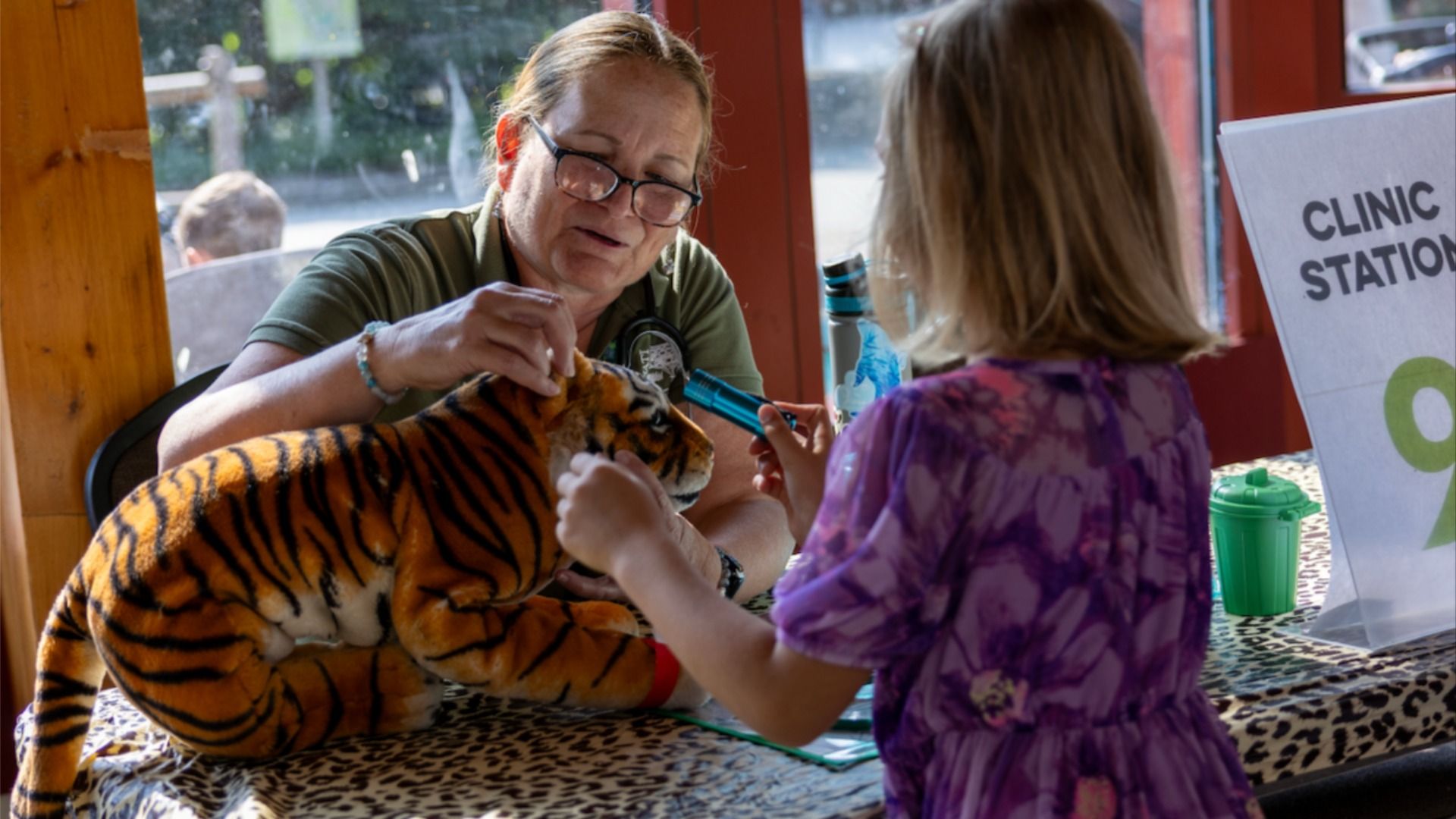 A woman with glasses and a stethoscope examines a tiger plush at a pretend clinic, as a child in a purple dress watches; a CLINIC STATION sign is visible.