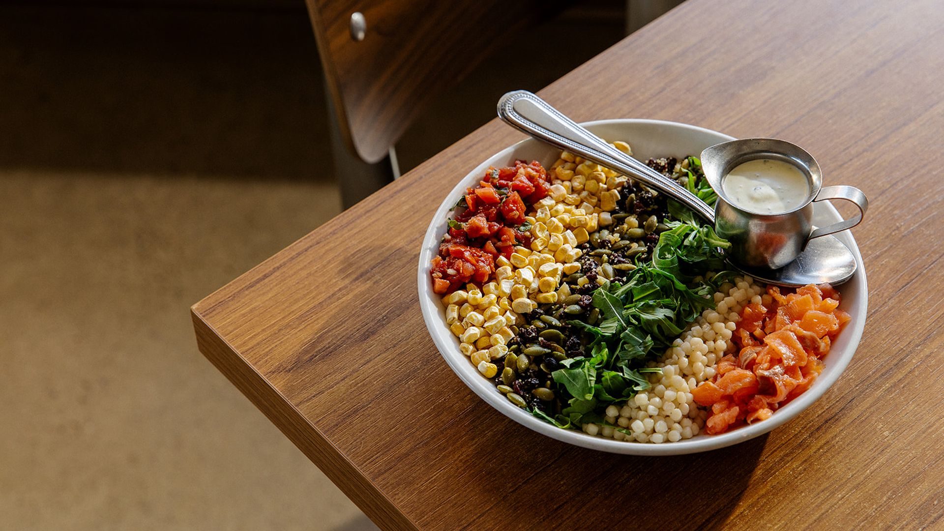 Large bowl of colorful mixed salad on a wooden table, featuring corn, diced tomatoes, greens, salmon chunks, and pearl couscous, with a metal pitcher of creamy dressing and a serving spoon.