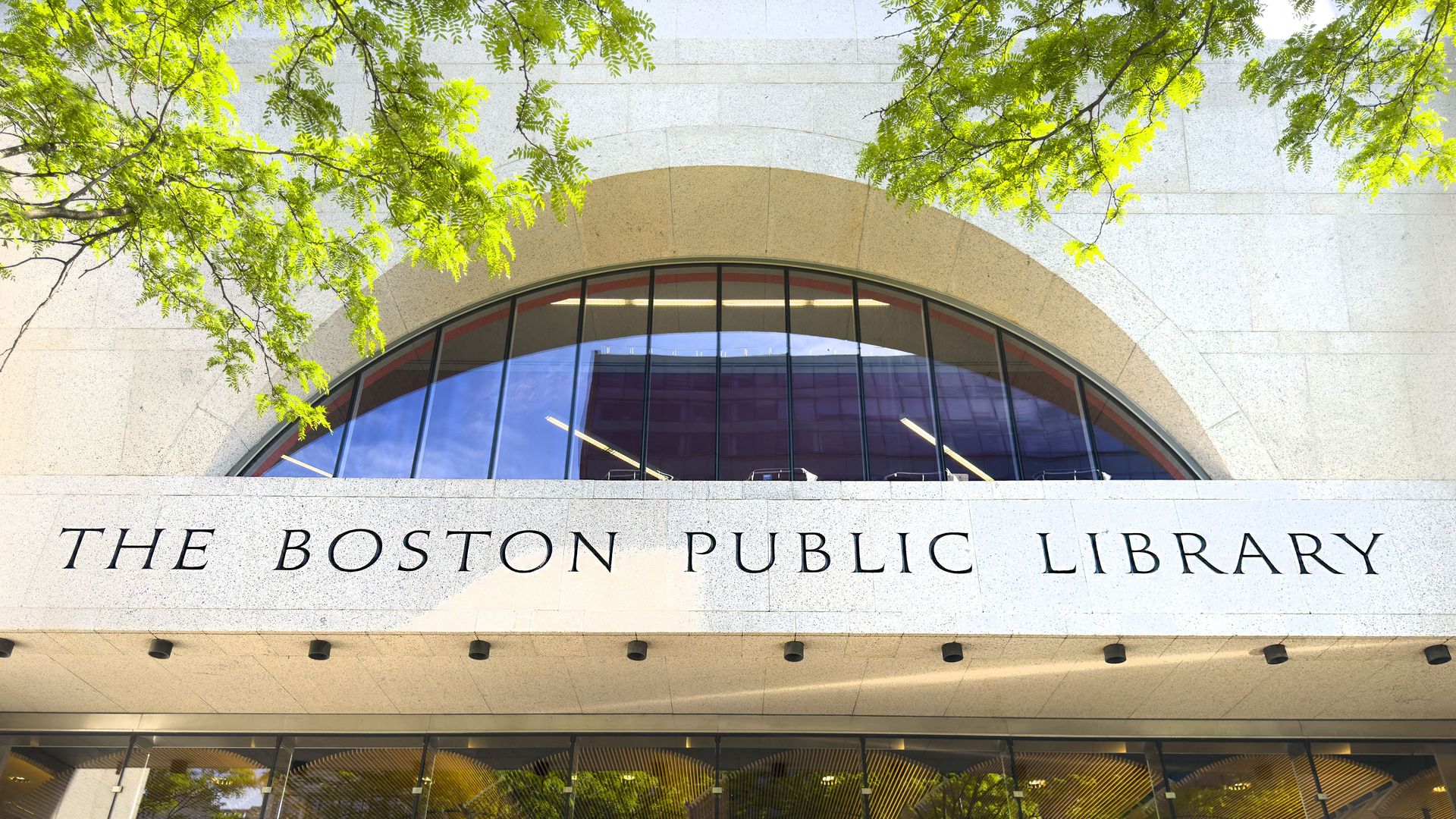 Front entrance of The Boston Public Library with large arched window and glass doors, framed by green tree branches and sunlight reflecting on the building.